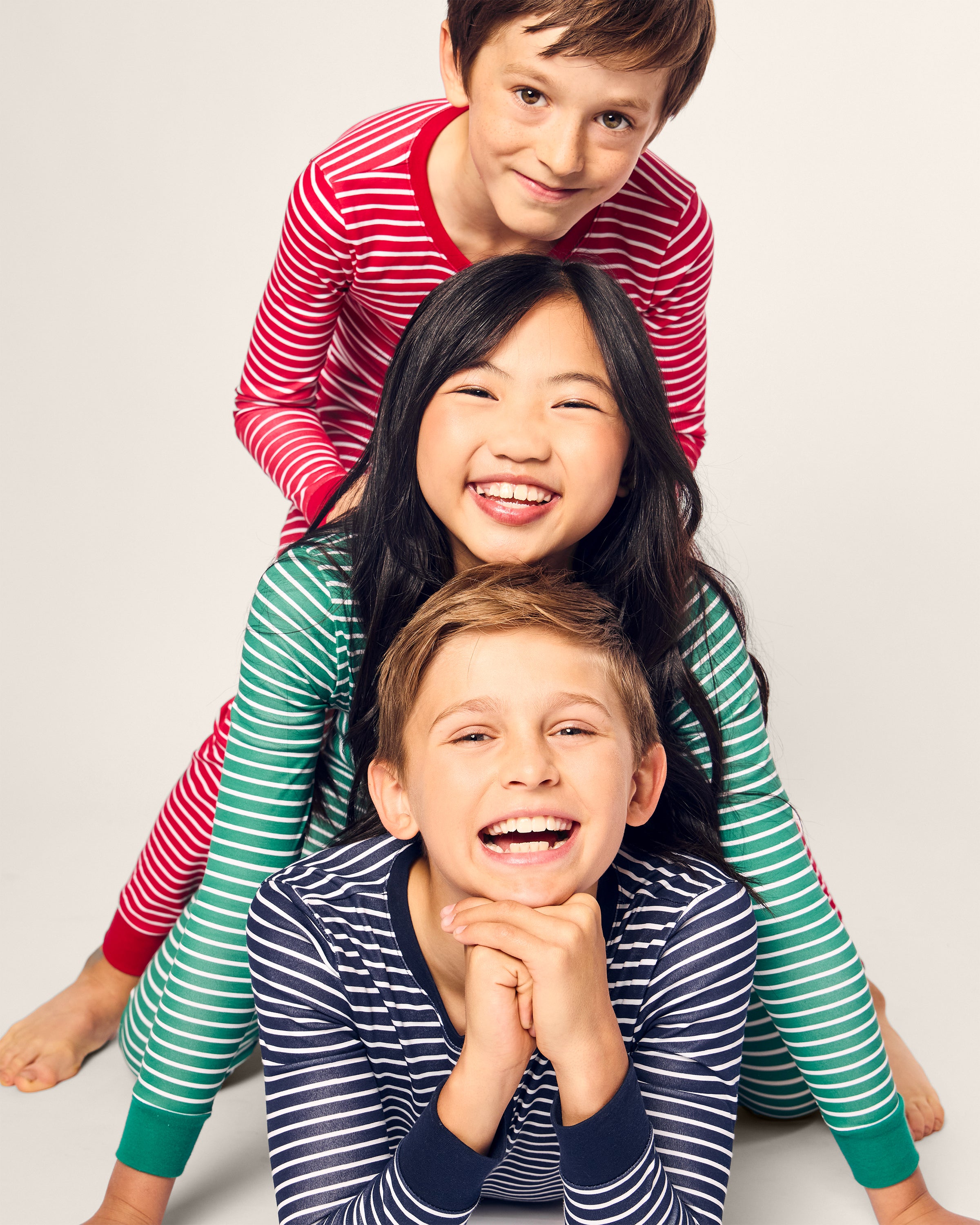 Three children in Petite Plumes Kids Pima Snug Fit Pajama Sets are playfully stacked, smiling. The bottom child in a blue set rests on their hands, the middle one in a green set grins widely, and the top child leans forward in a red stripe set. Theyre dressed in cozy long sleeves and pants.