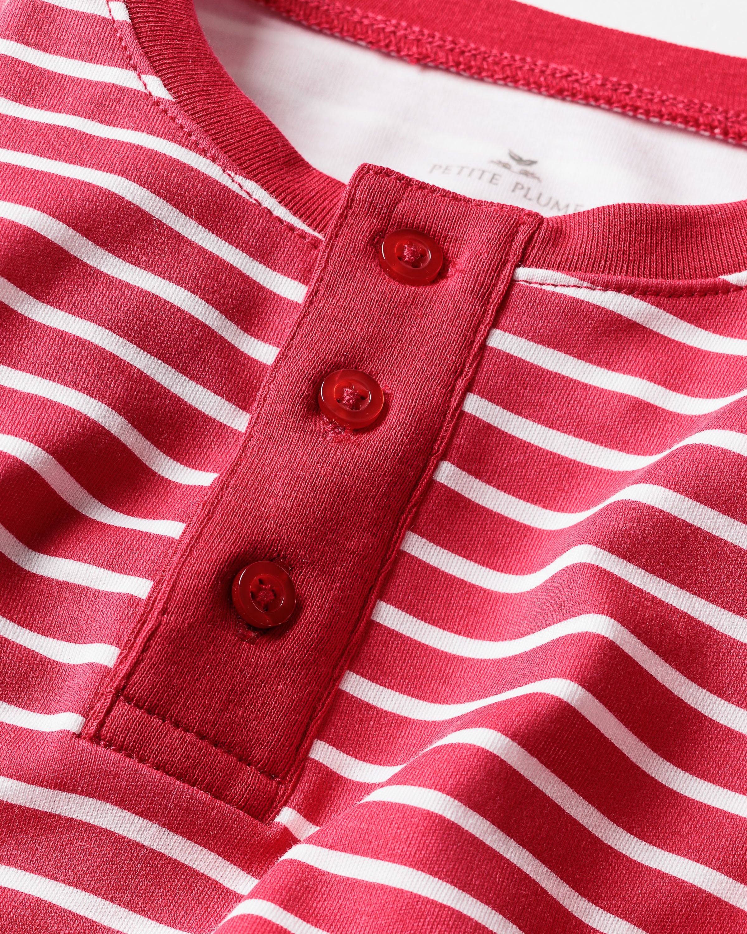 A close-up of a red and white striped shirt with a three-button placket showcases its Pima Cotton softness for a casual feel. The tag says Petite Plume, identifying it as the Kids Pima Snug Fit Pajama Set in Red Stripe.