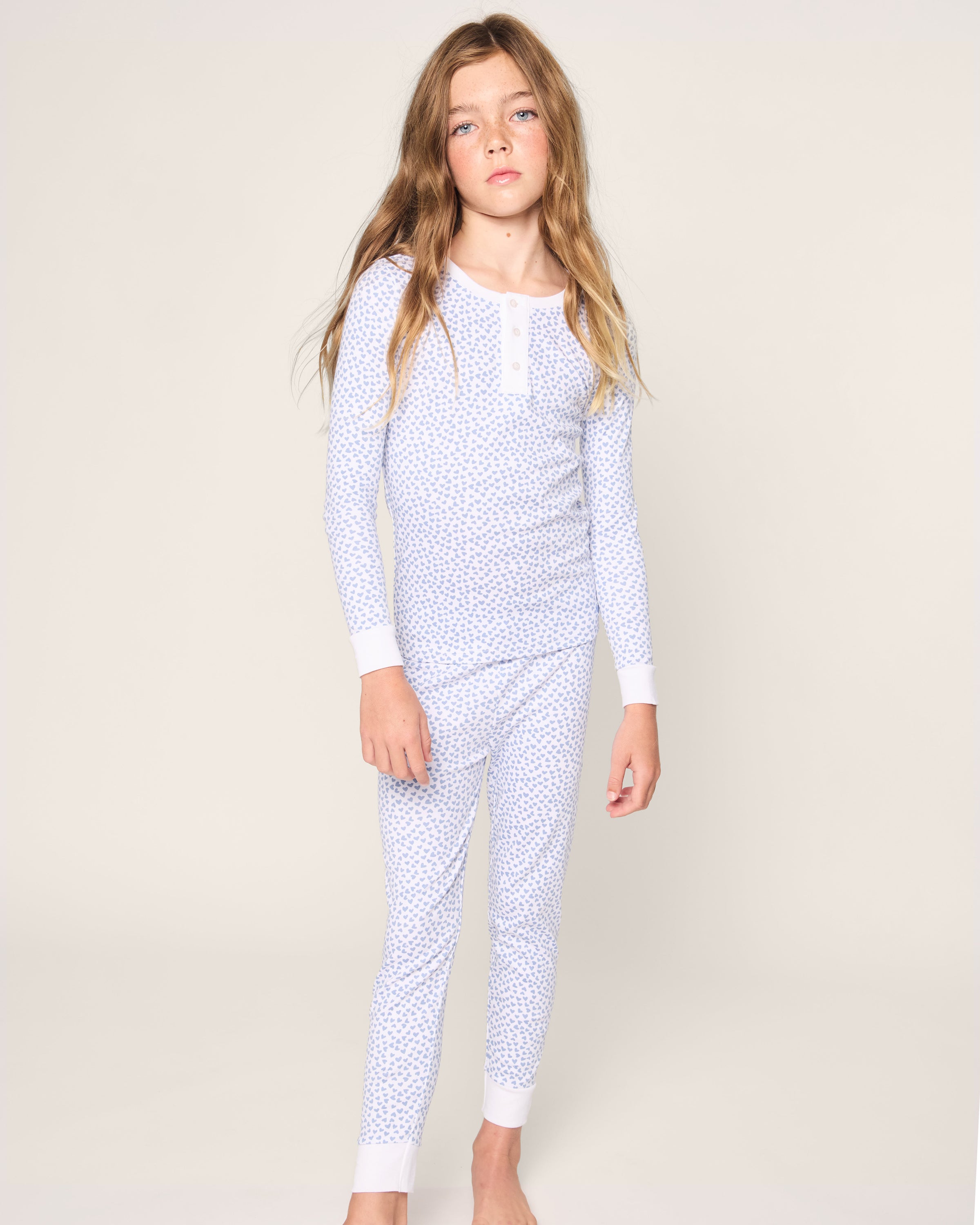 A young girl with long, brown hair poses against a light background in Petite Plumes Kids Pima Snug Fit Pajama Set in Bluehearts. The set features a soft, floral-patterned top and pants. Barefoot, she gazes at the camera with an innocent charm.