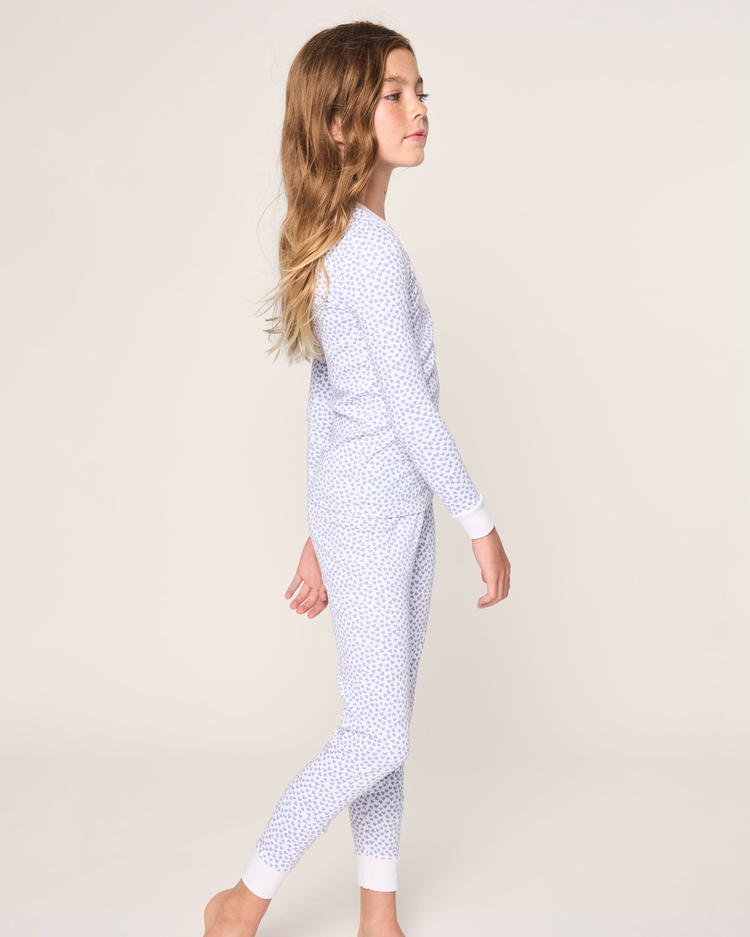 The girl in side profile wears a Petite Plume Kids Pima Snug Fit Pajama Set in Bluehearts, made from soft Pima cotton. Her long hair flows down naturally as she stands barefoot against a plain background.