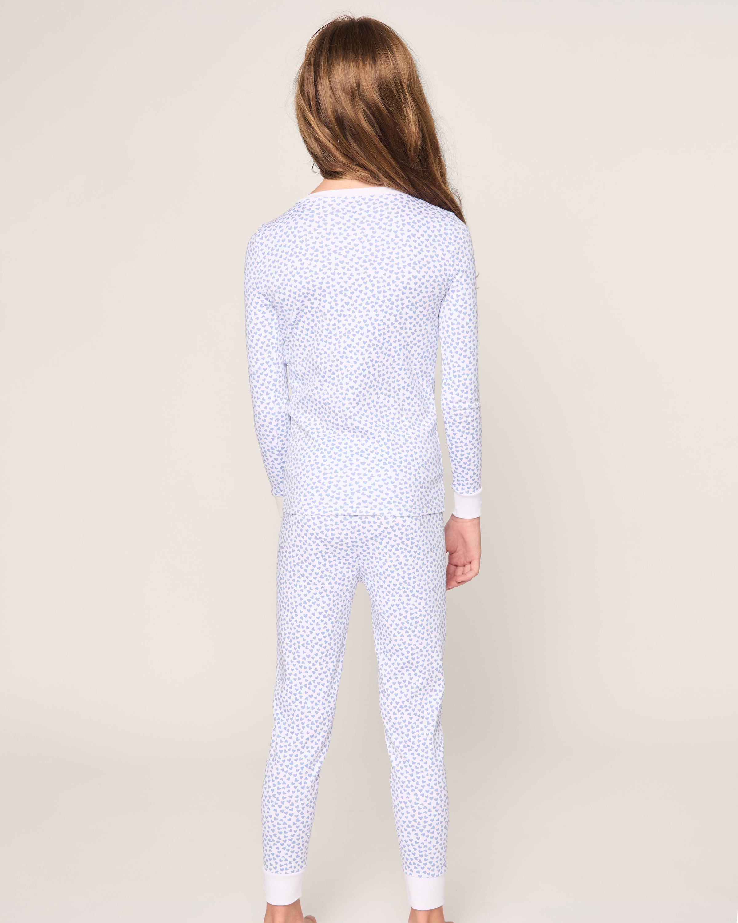 A child with long brown hair is seen from the back, donning the Kids Pima Snug Fit Pajama Set in Bluehearts by Petite Plume. The light-toned ensemble, adorned with small patterns and long sleeves, is made from soft Pima Cotton against an off-white background.