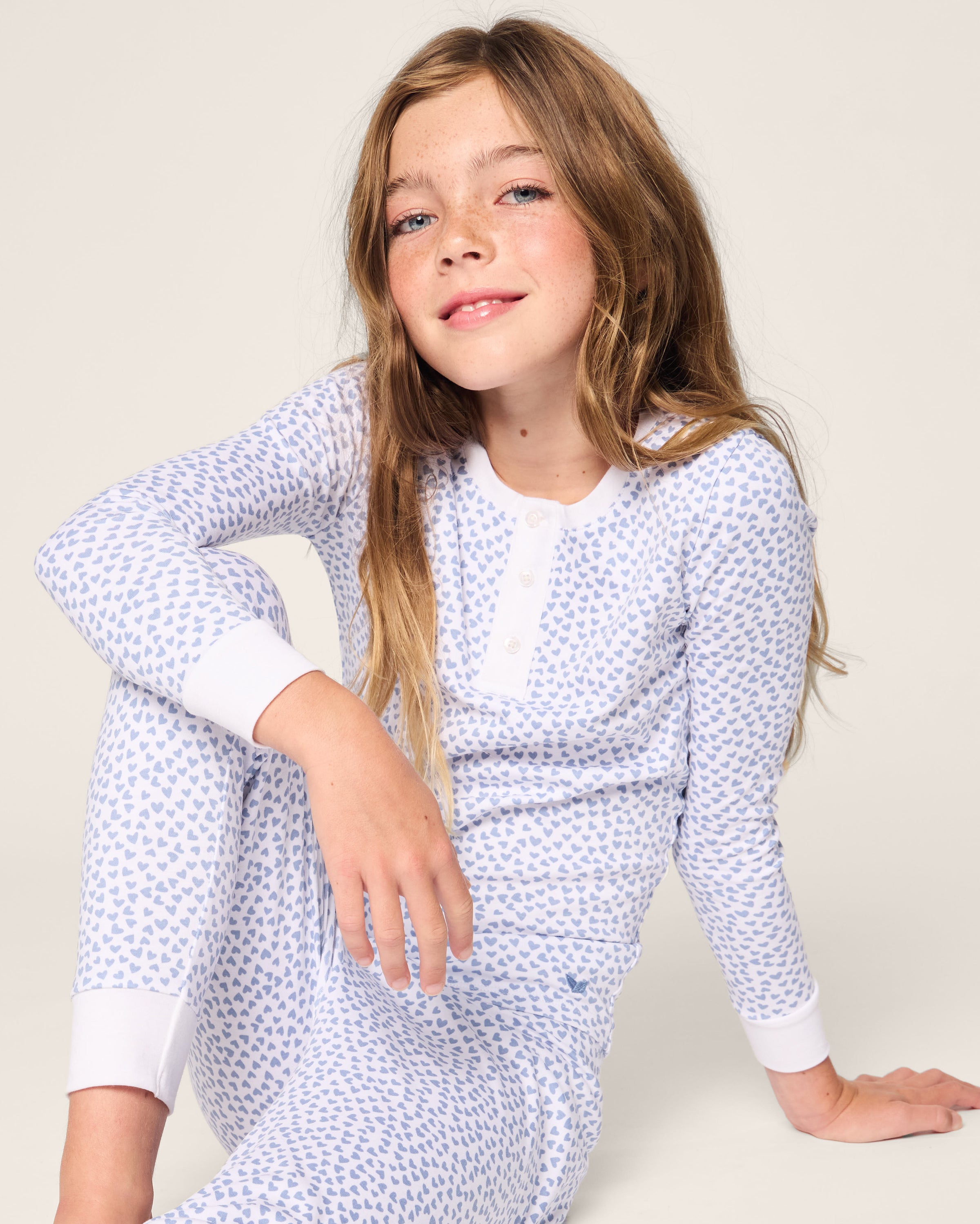 A young girl with long brown hair sits comfortably on the floor wearing a Petite Plume Kids Pima Snug Fit Pajama Set in Bluehearts. With one knee up, her soft smile enhances the sets cozy charm against a gentle background.