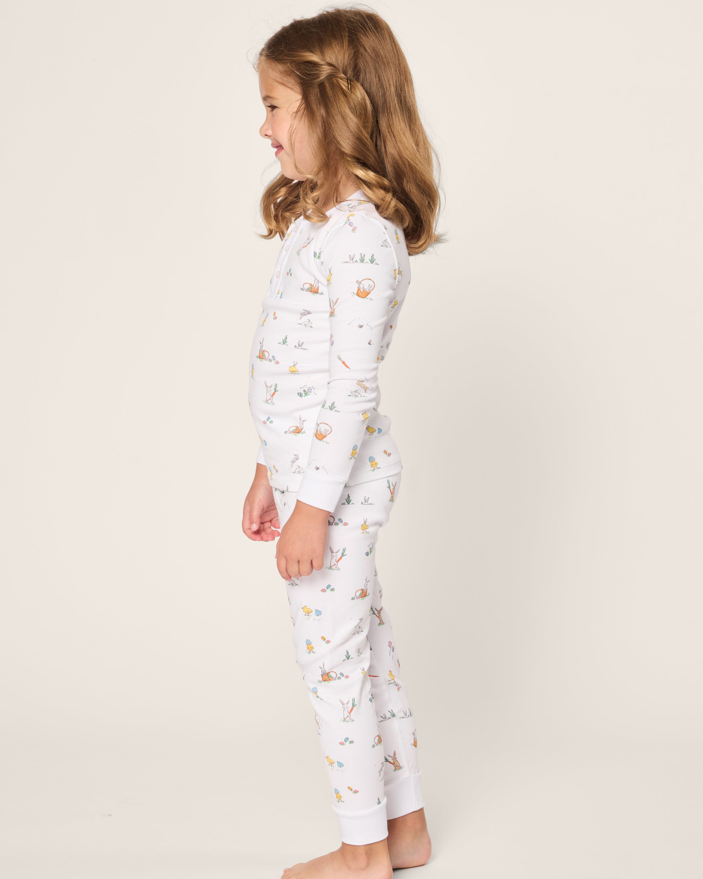 A child with long brown hair stands sideways in Petite Plumes Kids Pima Snug Fit Pajama Set in Easter Frolic, featuring a playful animal and plant pattern on soft white Pima Cotton, perfectly coordinated against a light background.