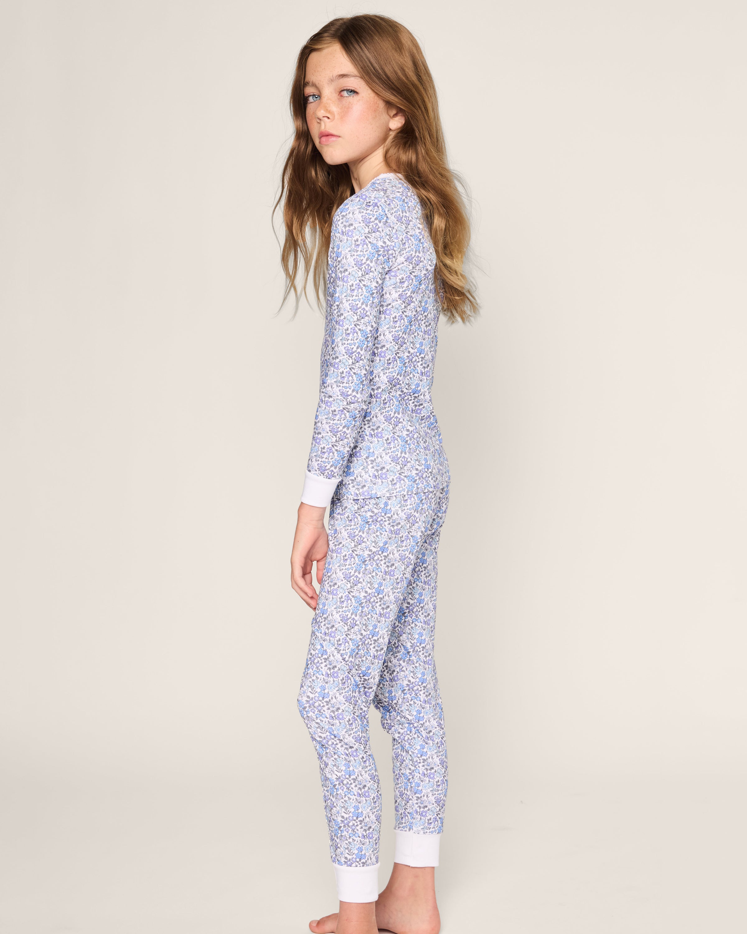 A young girl with long brown hair stands sideways, looking at the camera. She wears Petite Plumes Kids Pima Snug Fit Pajama Set in Fleur dAzur, featuring a cozy long sleeve top and pants made from soft Pima Cotton with a vintage-inspired blue and white floral print against a plain background.