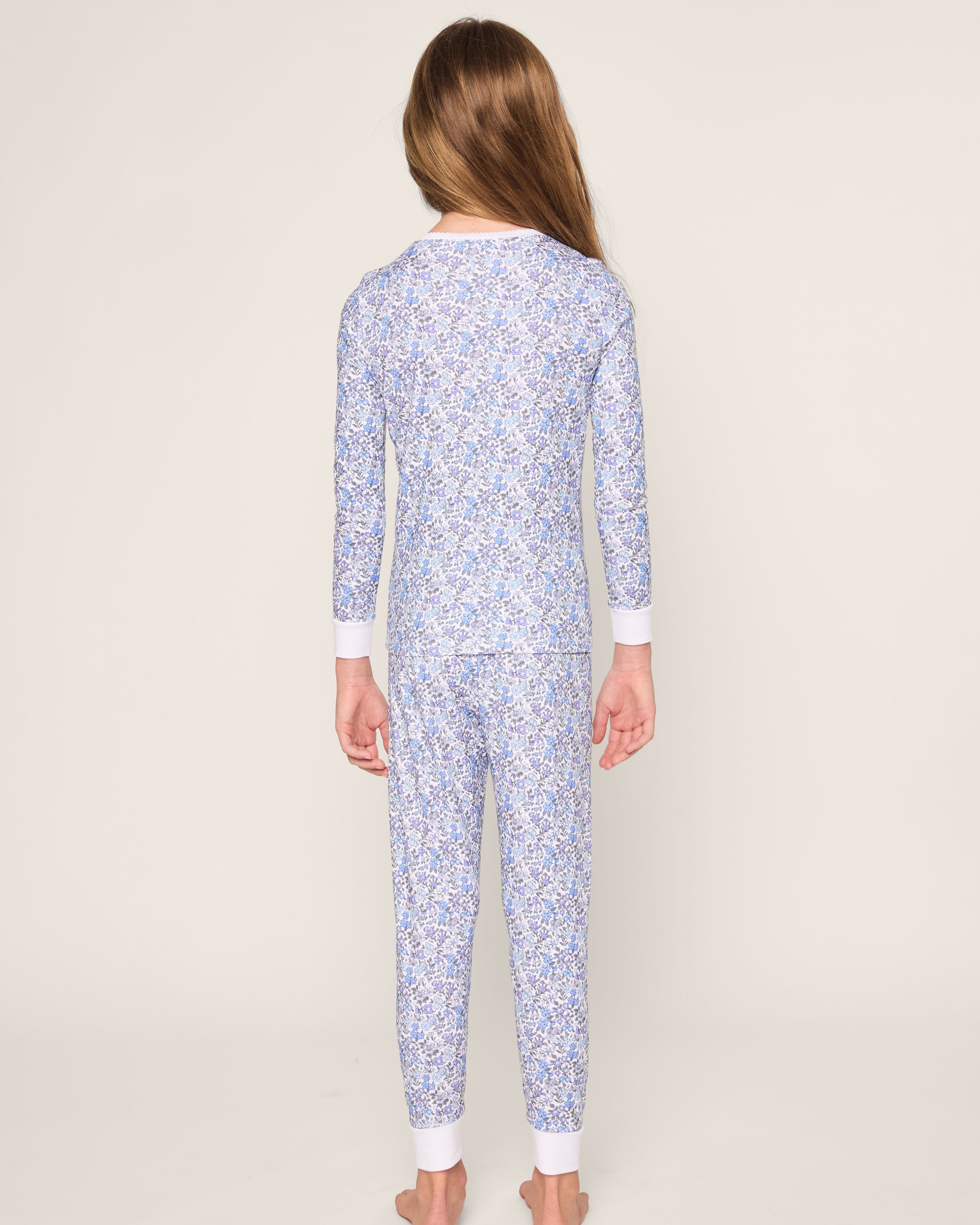 A child with long hair stands barefoot against a neutral background, wearing Petite Plumes Kids Pima Snug Fit Pajama Set in Fleur dAzur. The charming blue floral long sleeve top and pants set is crafted from soft Pima cotton, featuring a vintage-inspired print and white cuffs.