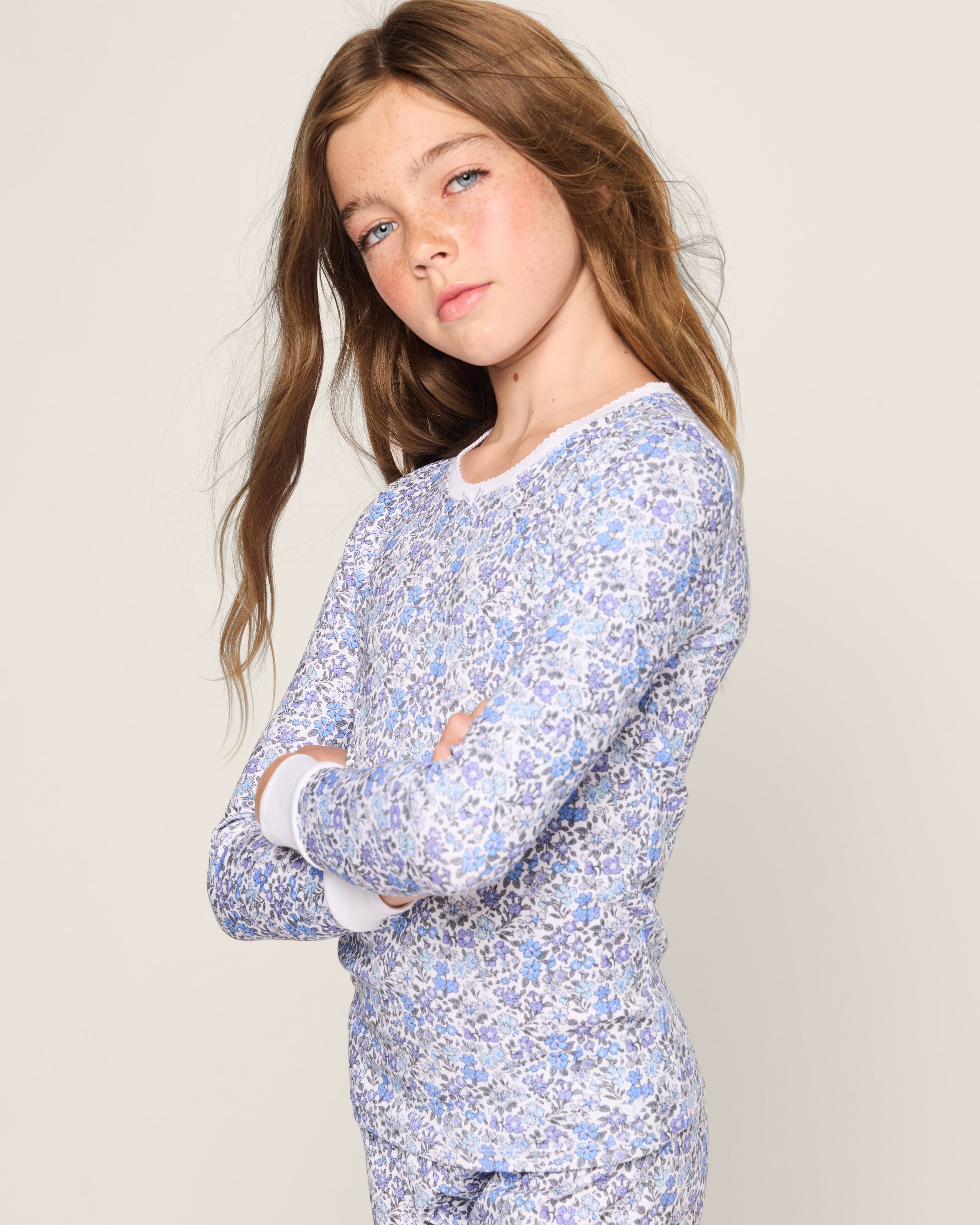 A young girl with long brown hair poses confidently, arms crossed, in Petite Plumes Kids Pima Snug Fit Pajama Set in Fleur dAzur. The vintage-inspired floral-patterned outfit crafted from soft Pima cotton stands out against the neutral background with timeless charm.