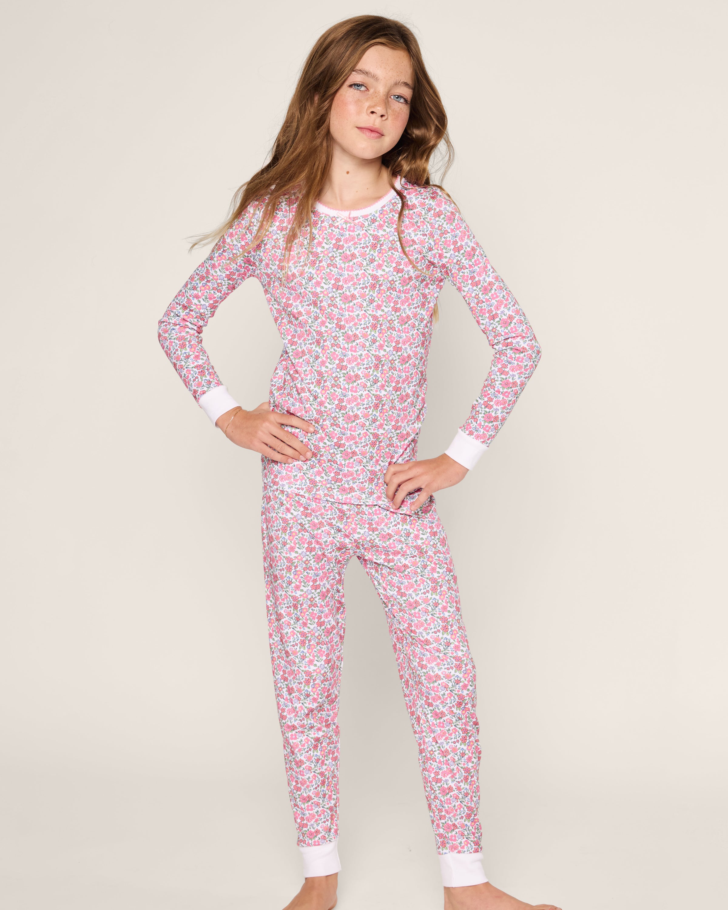 A young girl exudes confidence in Petite Plume’s Kids Pima Snug Fit Pajama Set in Fleurs de Rose print. The pink and white cotton sleepwear with ribbed cuffs complements her long brown hair and spirited expression against a plain background.