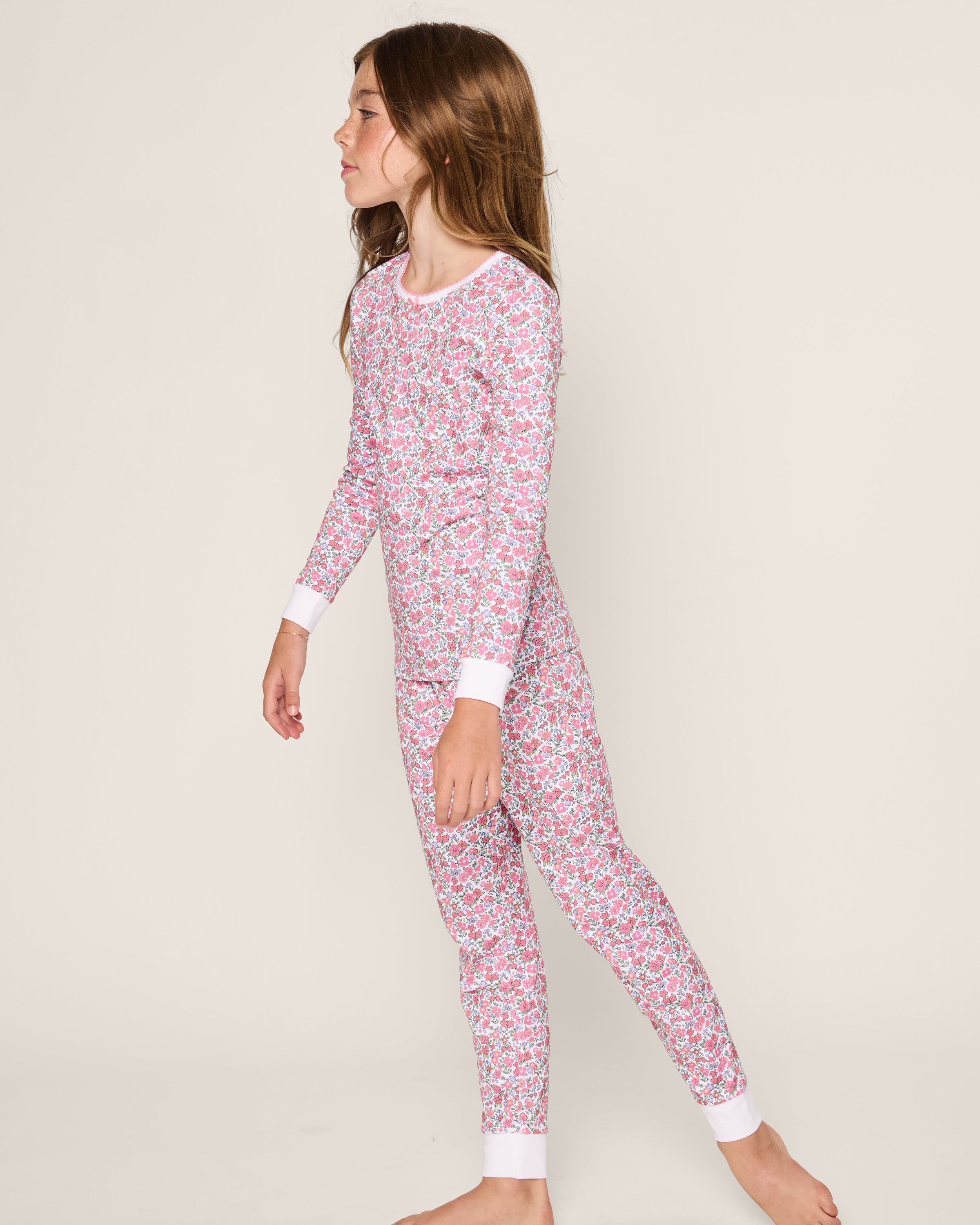 A young girl stands barefoot in a neutral backdrop, wearing Petite Plumes Kids Pima Snug Fit Pajama Set in Fleurs de Rose. The long-sleeve ensemble showcases charming pink and white hues of Pima Cotton as she gazes to the side.