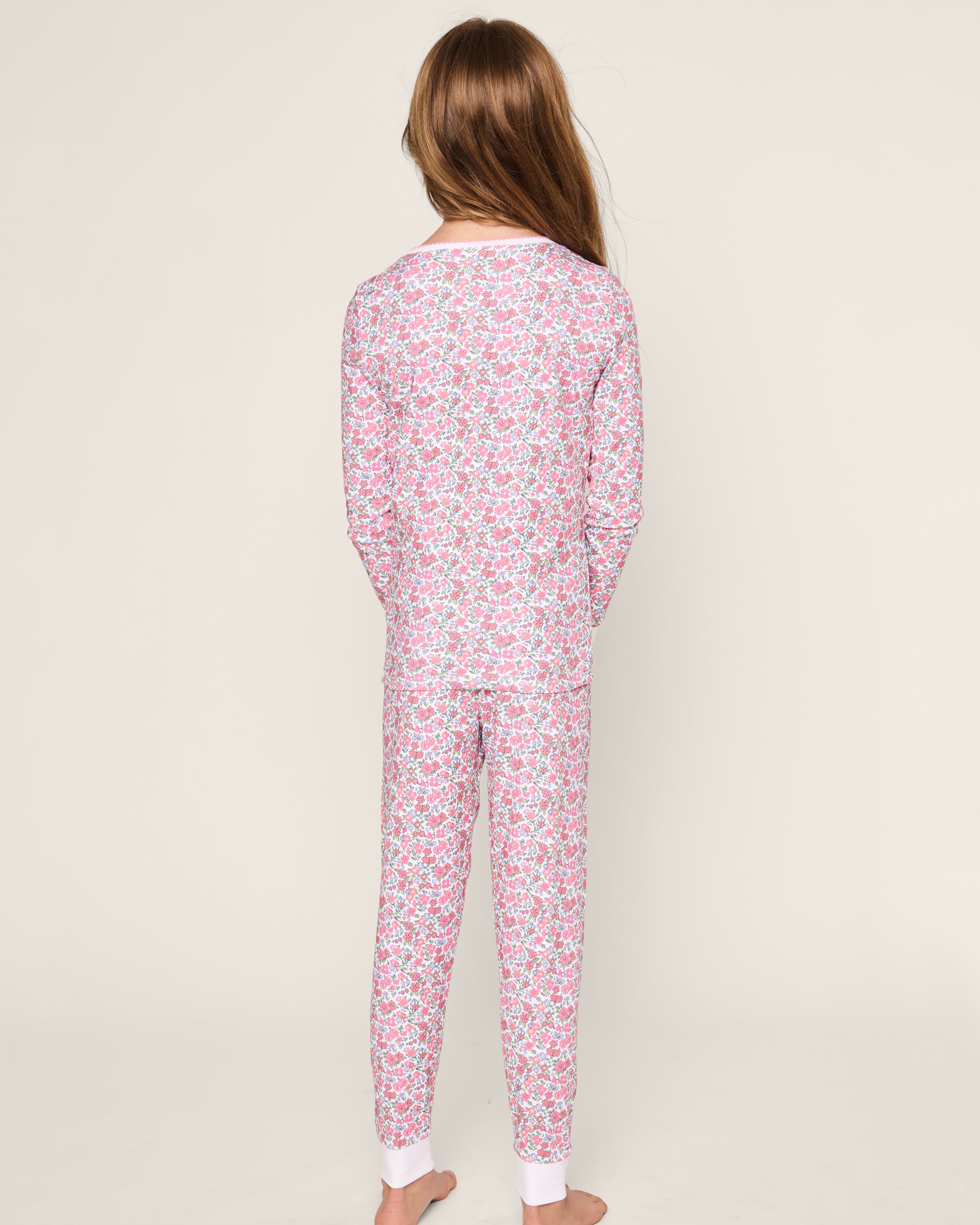 A child faces away, wearing Petite Plumes Kids Pima Snug Fit Pajama Set in Fleurs de Rose, a floral-patterned onesie in pink, purple, and white tones. The background is plain off-white.