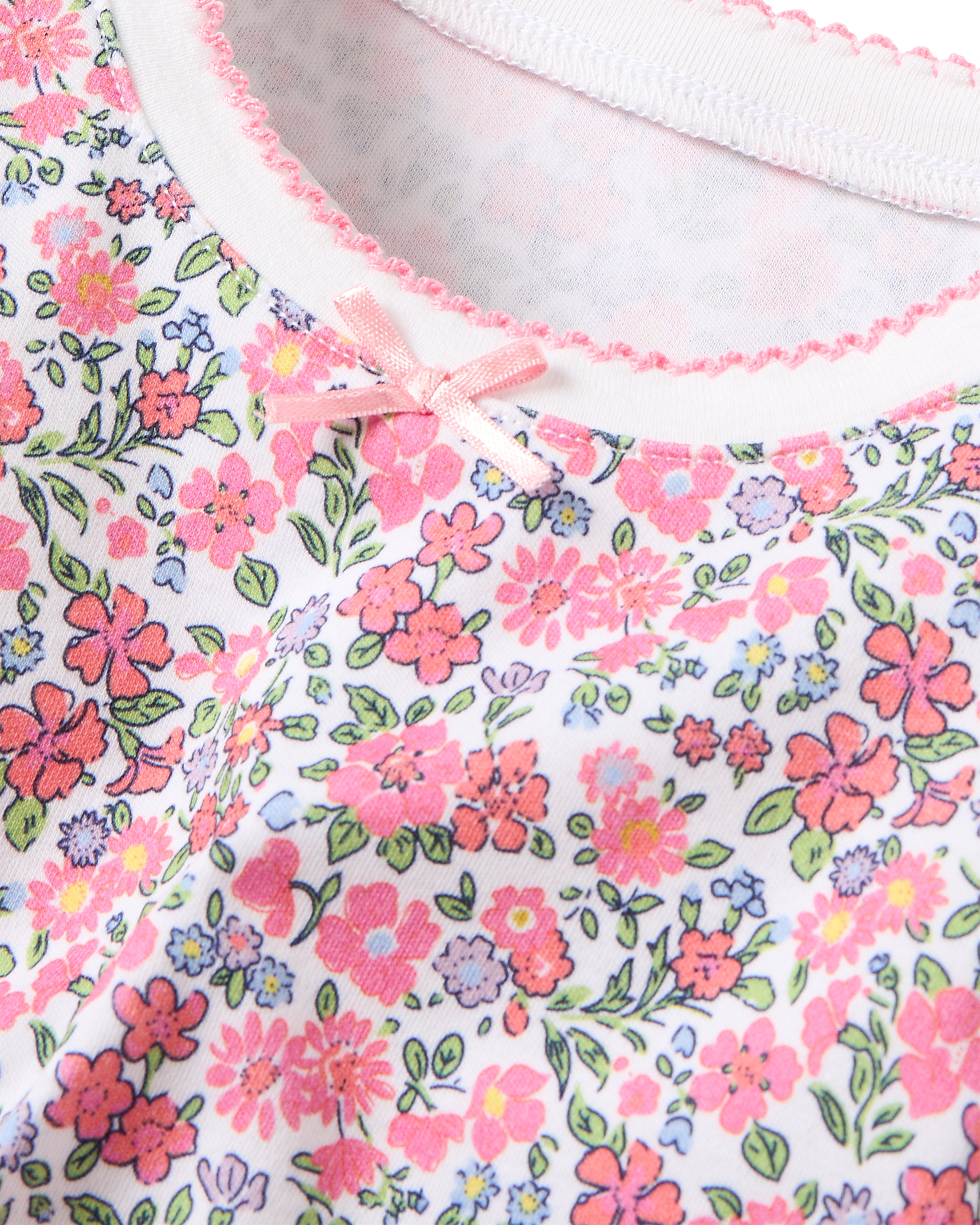 Close-up of Petite Plumes Kids Pima Snug Fit Pajama Set in Fleurs de Rose, highlighting pink, red, and purple floral print with green leaves on white. It features a snug fit design with a pink bow and scalloped trim along the neckline, embodying the comfort of Pima Cotton Sleepwear.