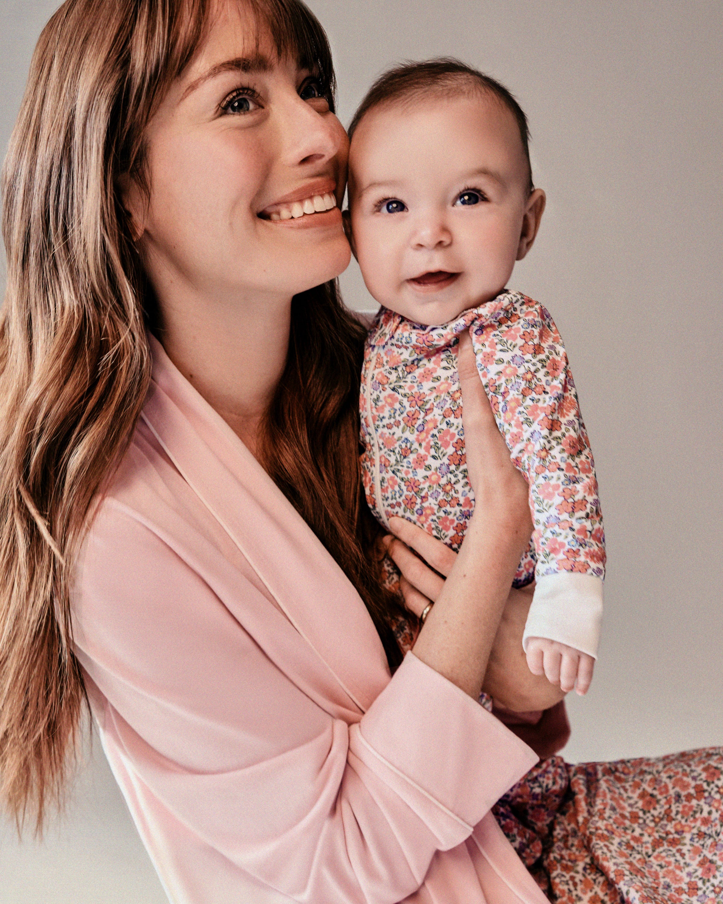 A woman, smiling, holds a baby in a charming Petite Plume Pima Ruffle Collar Romper in Fleurs de Rose print. The woman wears a vibrant pink robe. Both look happy and content against a neutral background.