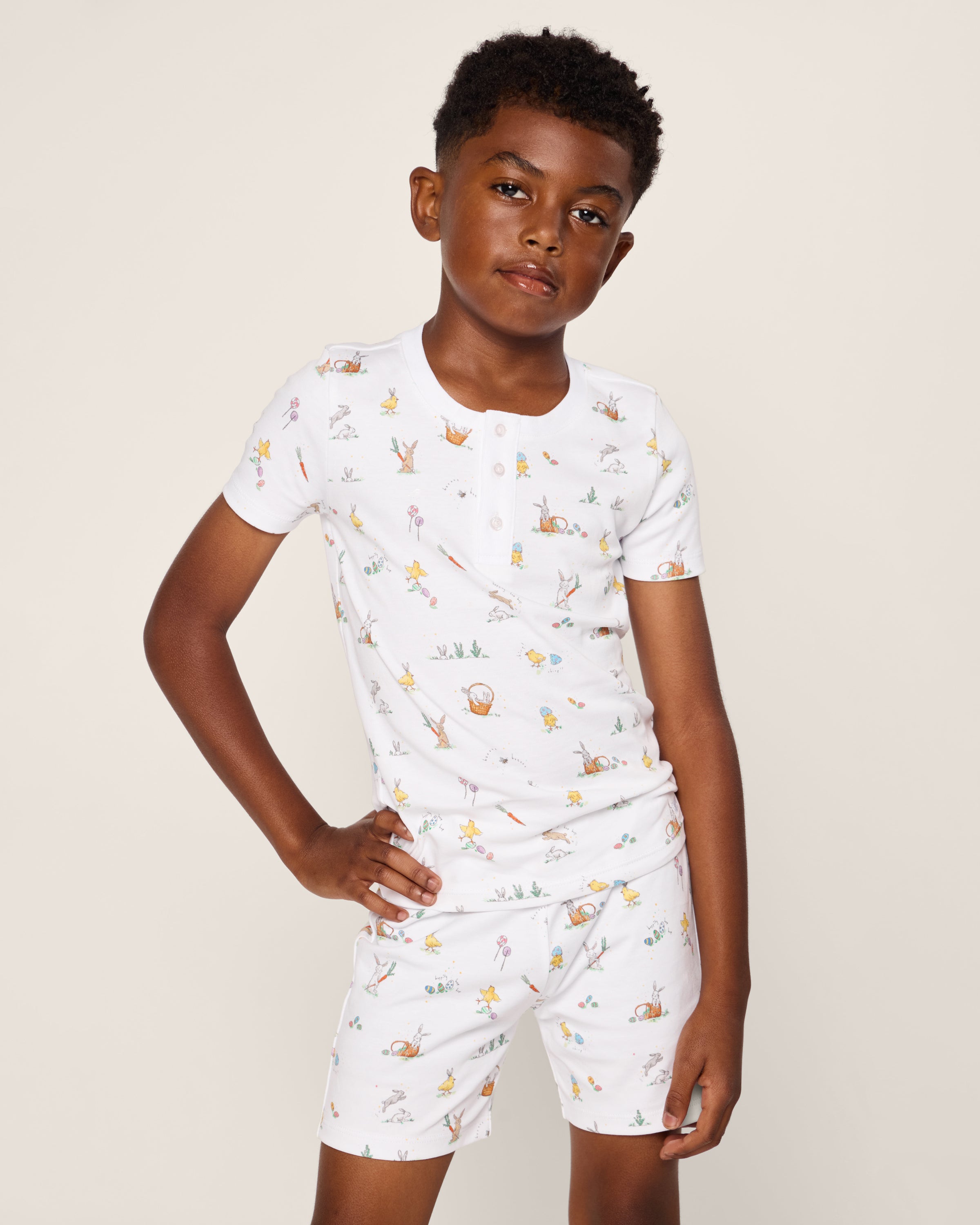 A child stands confidently with one hand on their hip, wearing Petite Plumes Kids Pima Snug Fit Short Set in Easter Frolic, featuring luxurious white Pima Cotton with a colorful animal and nature print. The background is a plain, light-colored wall.