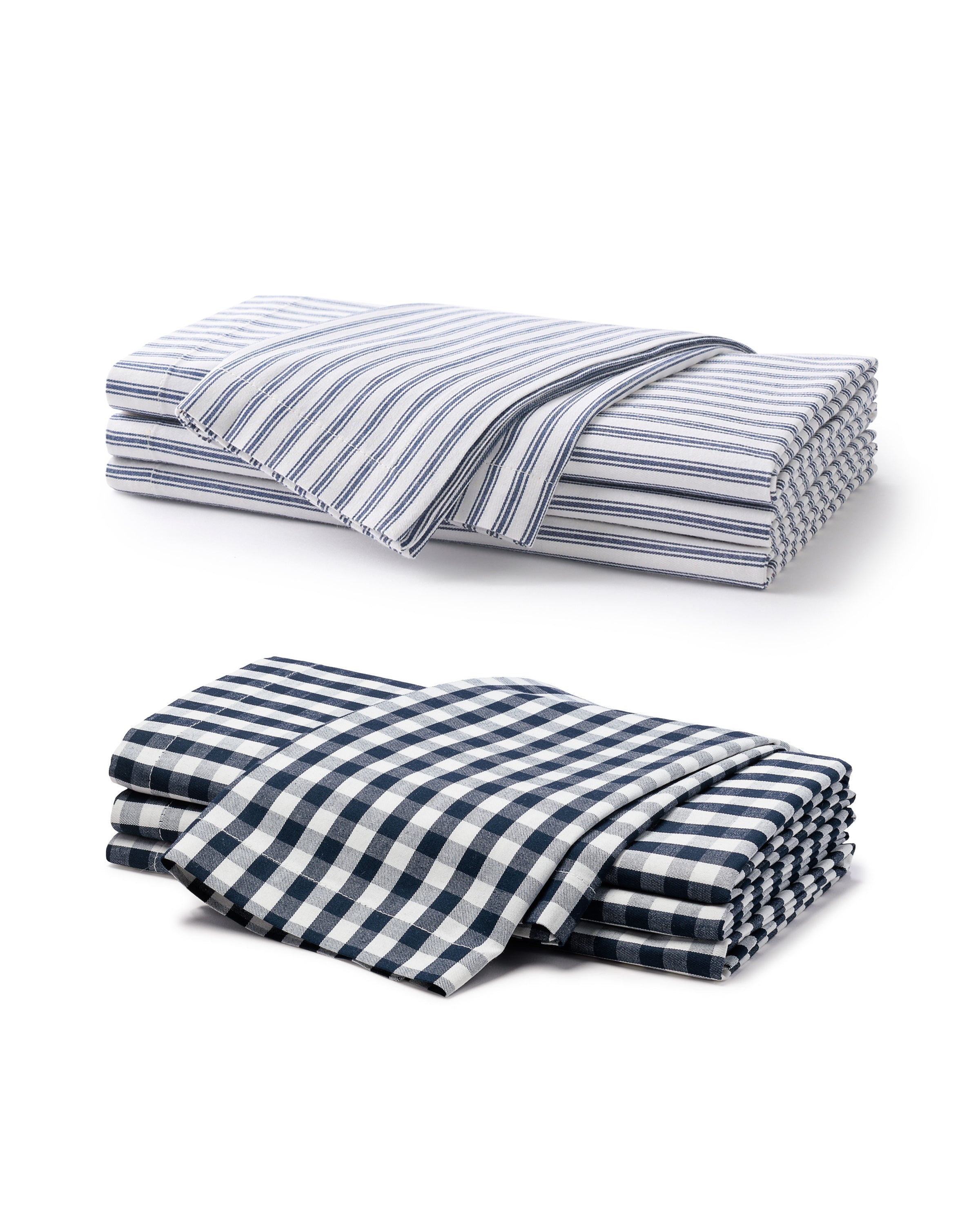 Elegantly layered in the image, a stack of Petite Plumes Signature Twill Napkin Set showcases a white and blue pinstripe pattern on top and a navy and white gingham design below. Crafted from 100% cotton, these table linens feature classic prints against a crisp white background.