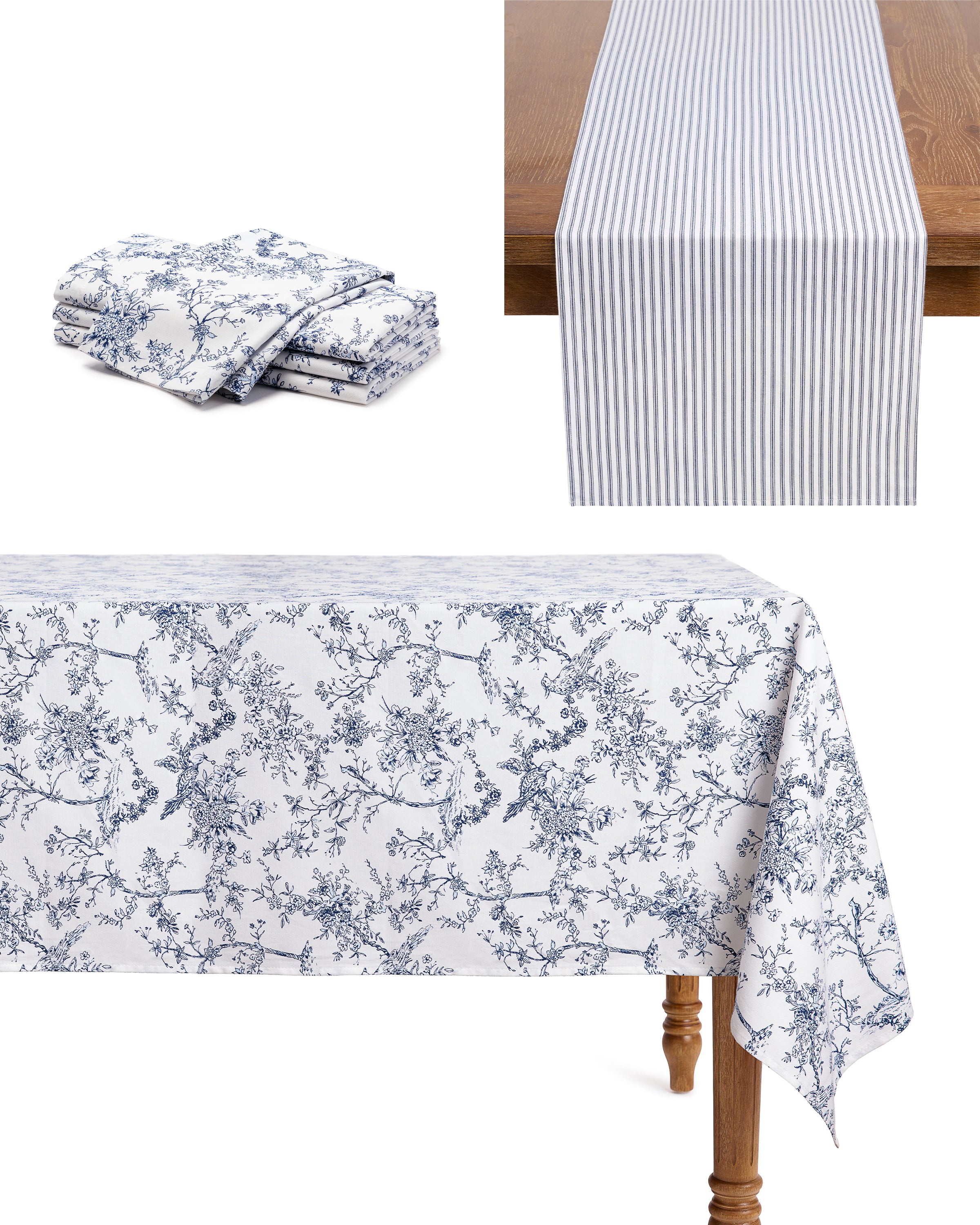 A table is decorated with Petite Plumes Signature Twill Table Linen set in Timeless Toile & Navy French Ticking, featuring a white cloth with a blue floral pattern and matching folded items. A runner with Petite Plume designs in blue and white stripes adds elegance to the wooden tablescape.