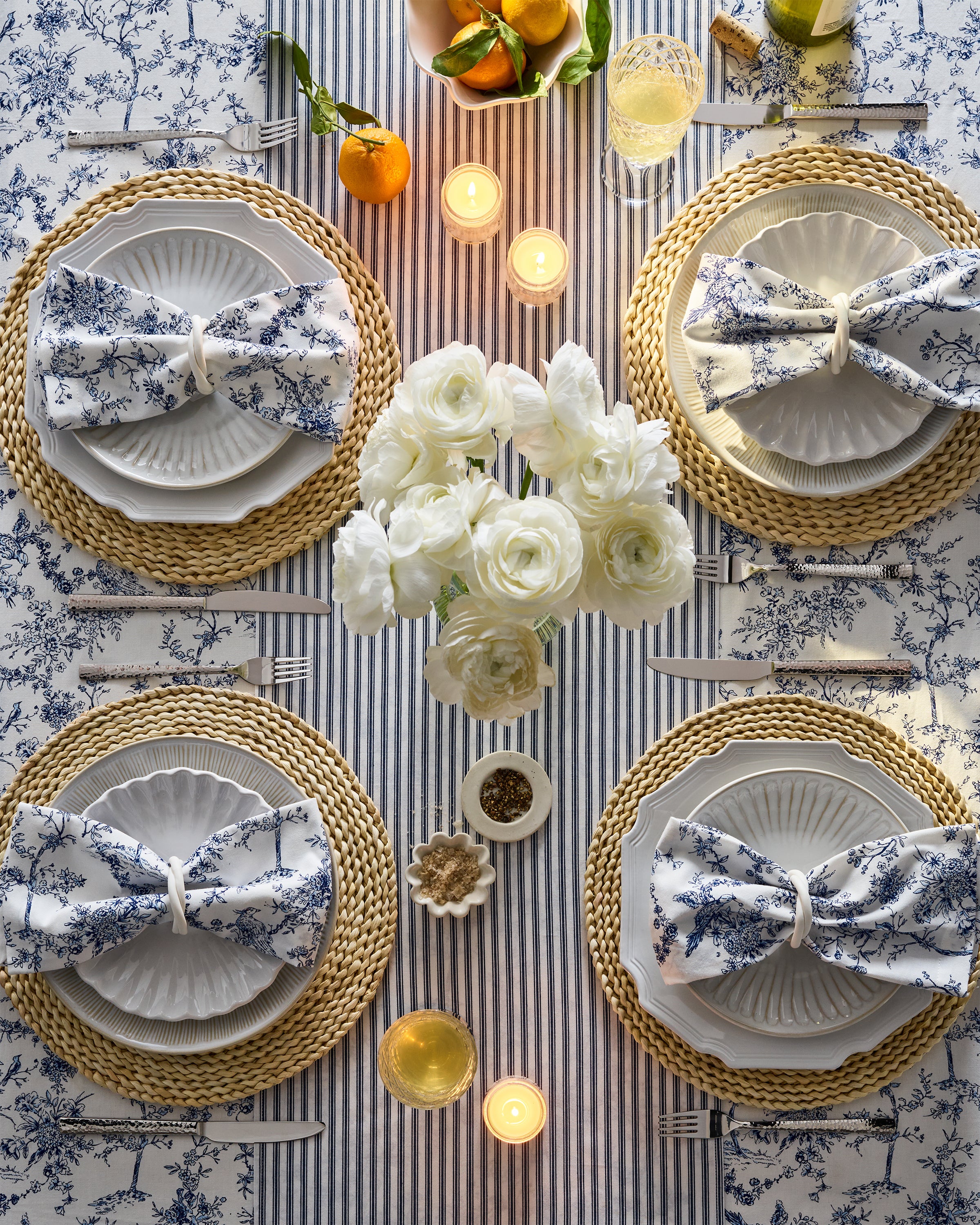 A top view of a dining table set with Petite Plumes Signature Twill Table Linen in Timeless Toile & Navy French Ticking, floral napkins on plates, woven placemats, white flowers, lit candles, and citrus fruits creates an elegant and cozy festive tablescape.