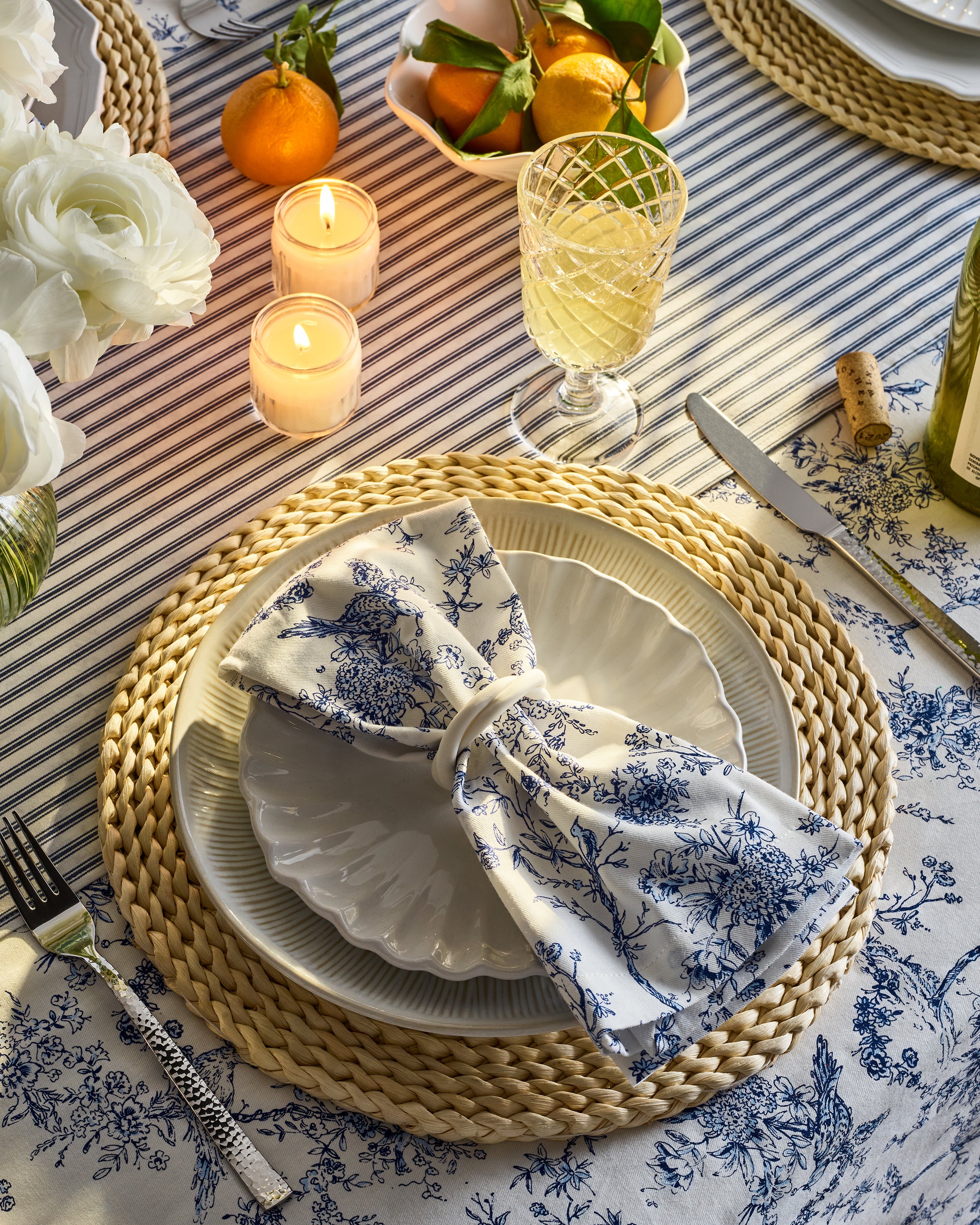 A beautifully set table with a floral-patterned napkin tied into a bow on a white plate graces the festive tablescape. Surrounded by candles, oranges, and white wine, it rests upon Signature Twill Table Linen in Timeless Toile & Navy French Ticking by Petite Plume.