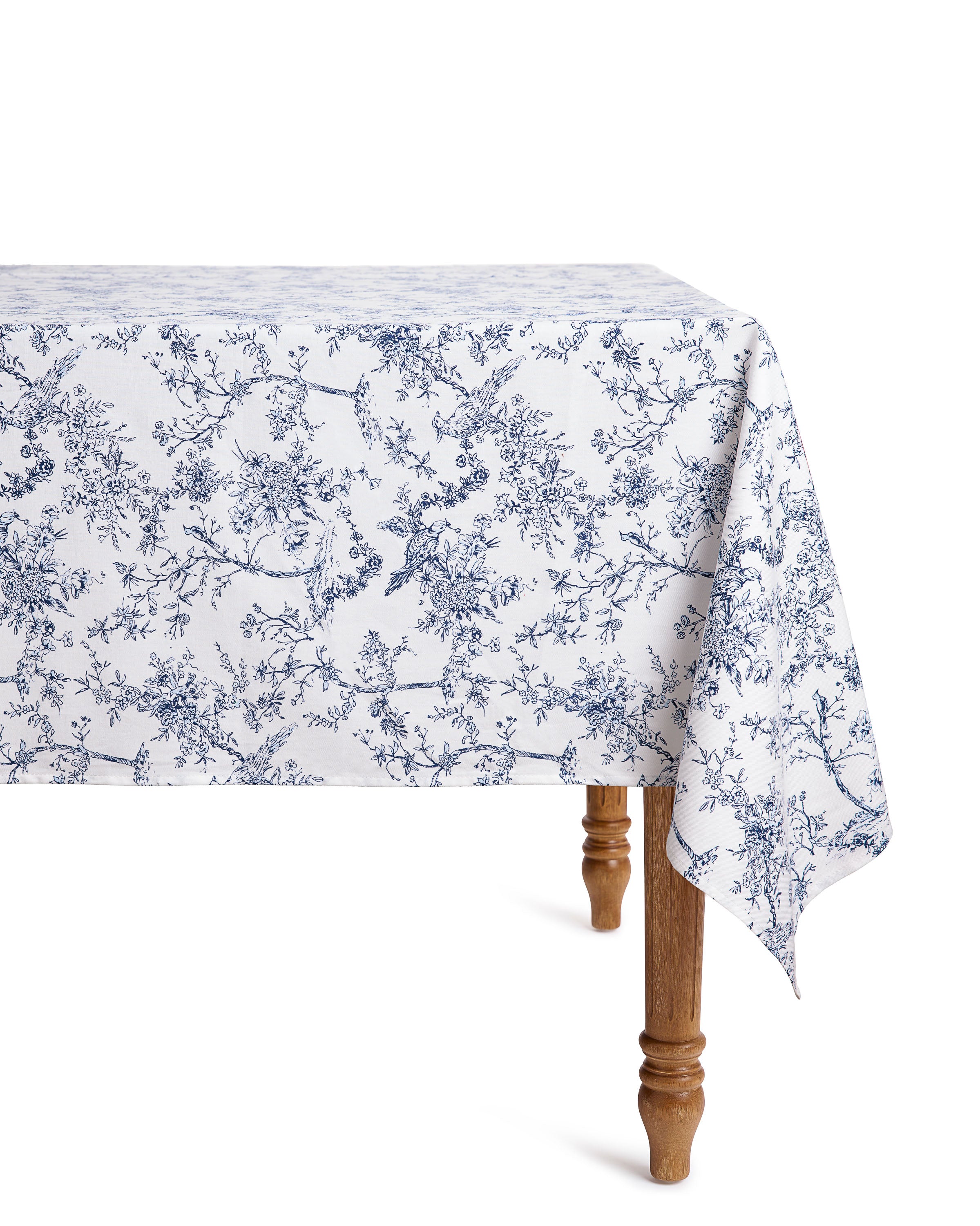 A wooden table is set with Petite Plumes Signature Twill Table Linen set in Timeless Toile & Navy French Ticking, featuring a blue floral pattern of branches and flowers. The linens elegantly drape over the edge, crafting a festive tablescape, with one of the tables legs visible.