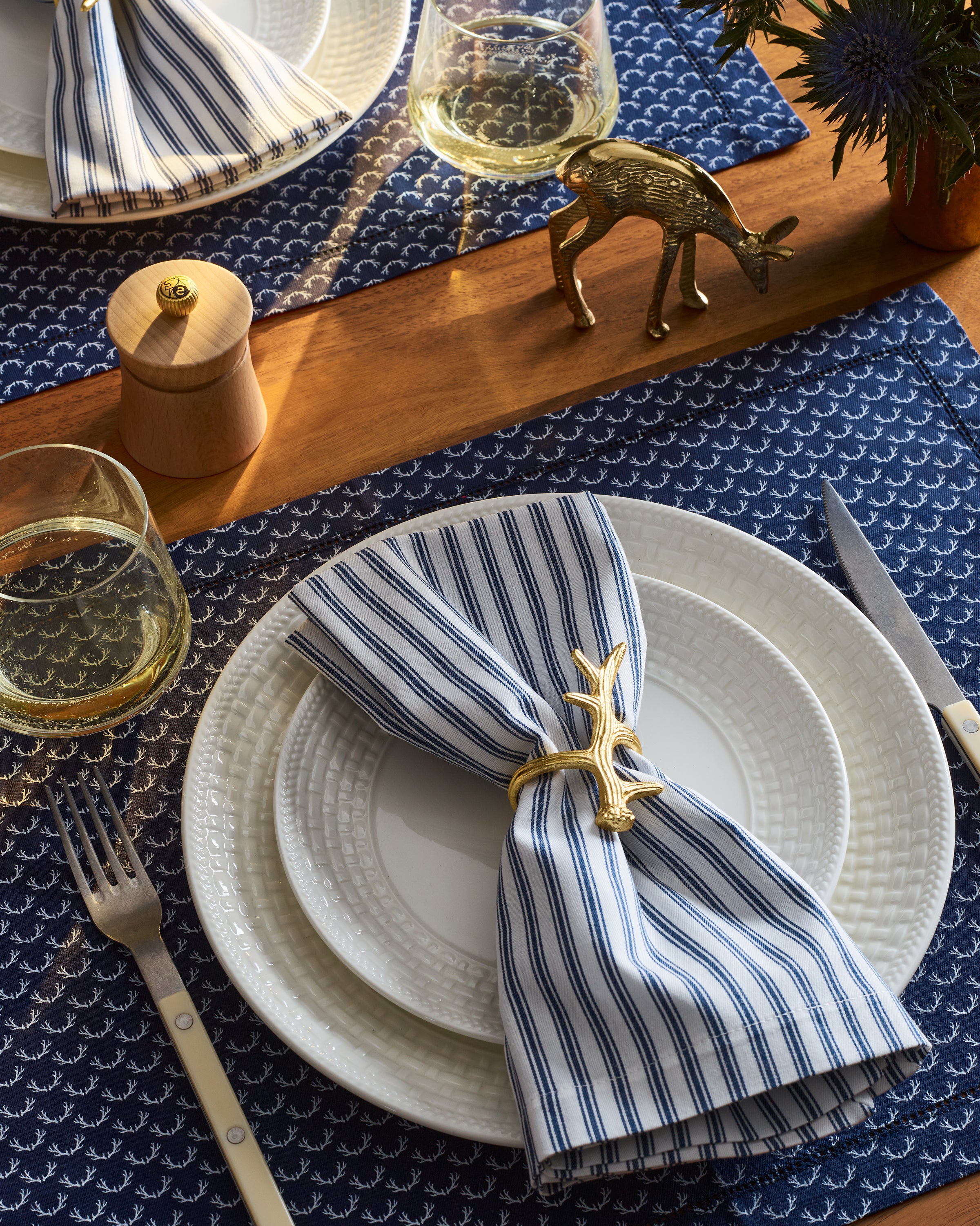 A festive tablescape awaits with an elegant dining set featuring white plates, Petite Plumes Signature Twill Napkin in Navy French Ticking secured by golden napkin rings. Wine glasses and silverware complement a small brass deer ornament on blue placemats with a white pattern.