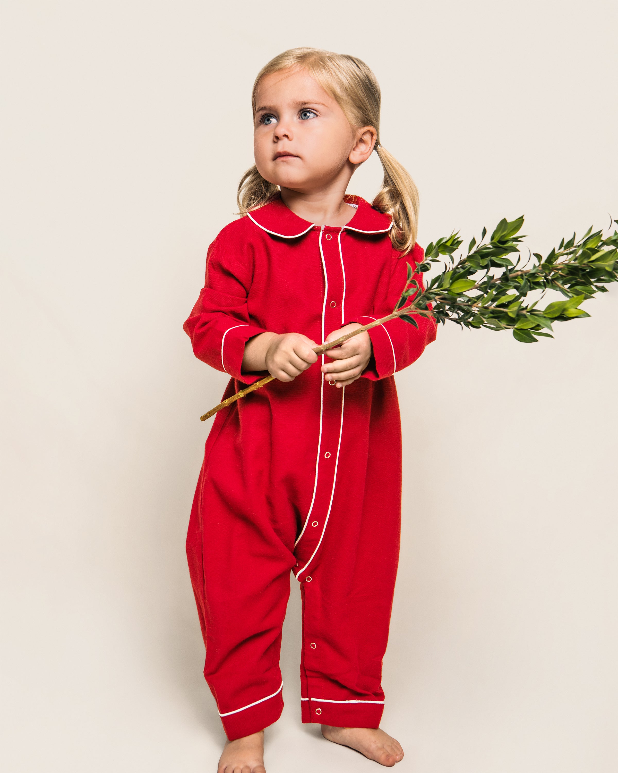 A young child with blonde pigtails stands barefoot, holding a branch with green leaves. They are wearing a red Flannel Cambridge Romper with white trim by Petite Plume against a plain backdrop, resembling an adorable part of the Babys Flannel Cambridge Romper & Cashmere Blanket Gift Set.