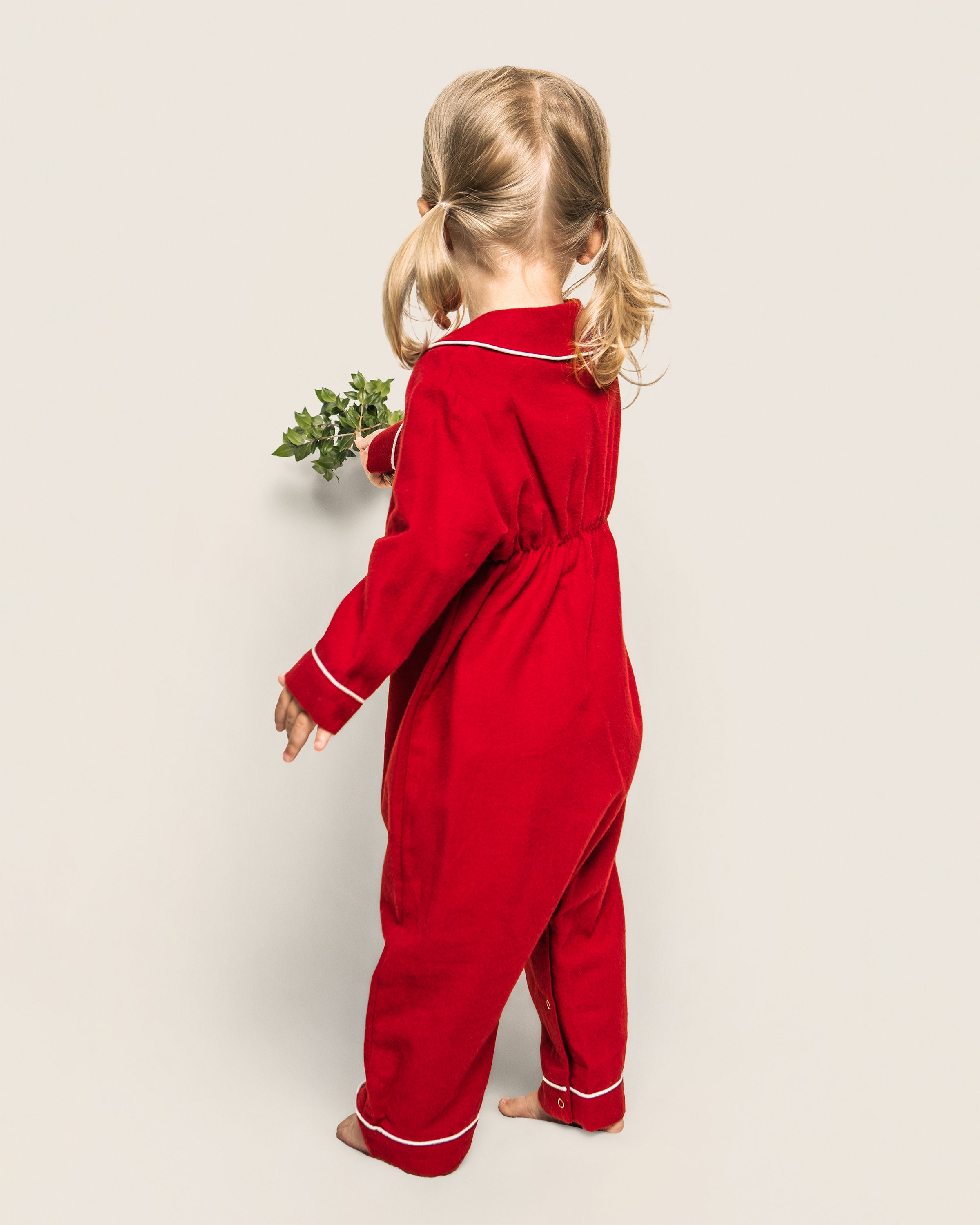 A small child with blond pigtails is wearing a red onesie from Petite Plumes Babys Flannel Cambridge Romper & Cashmere Blanket Gift Set, standing barefoot holding a small green plant against a plain background, facing away.