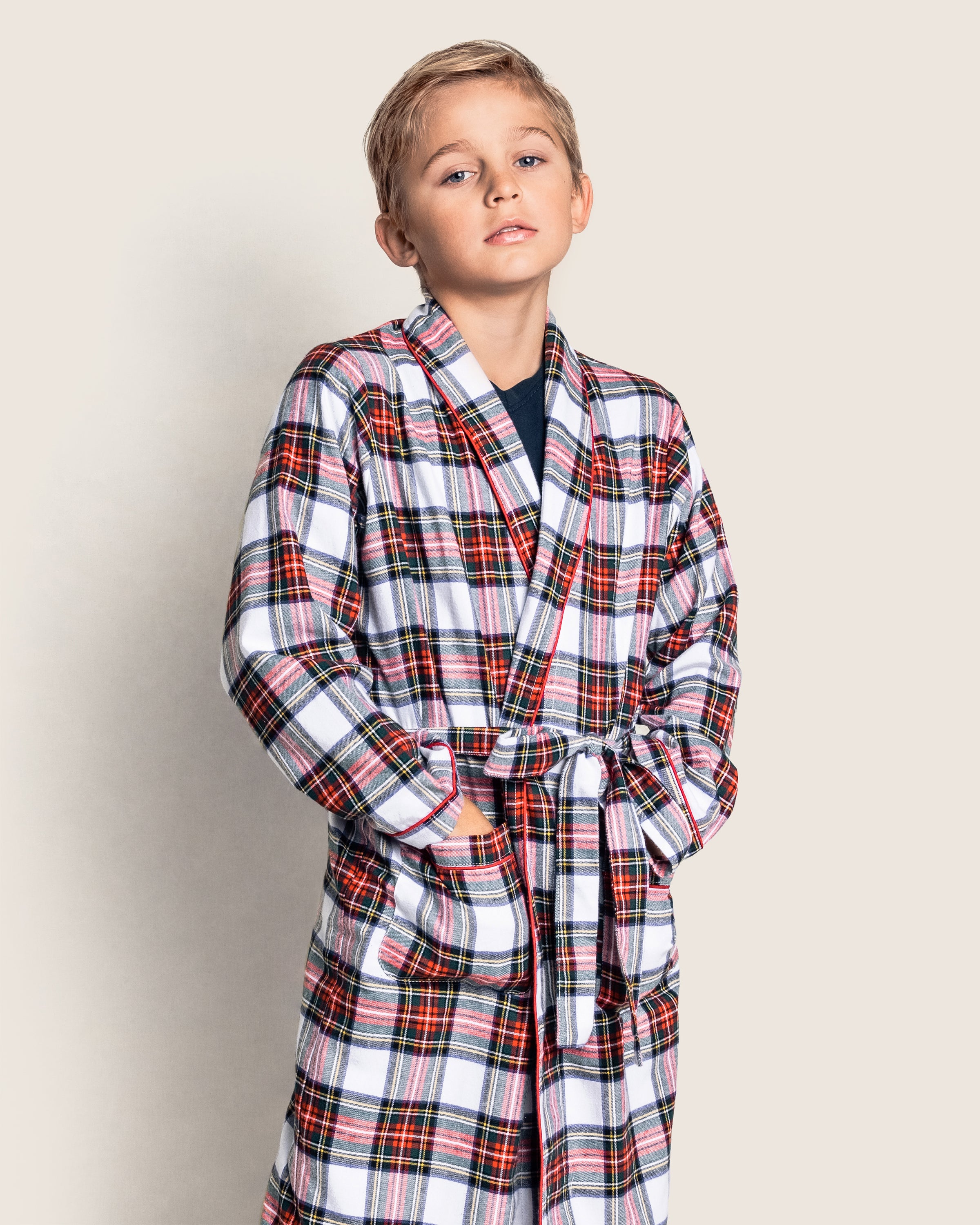 The boy stands confidently in his red and white Petite Plume Kids Brushed Cotton Robe in Balmoral Tartan, hands tucked into pockets. Made from flame-retardant fabric, this stylish sleepwear keeps him safe as he gazes at the camera with a slight head tilt against a plain backdrop.