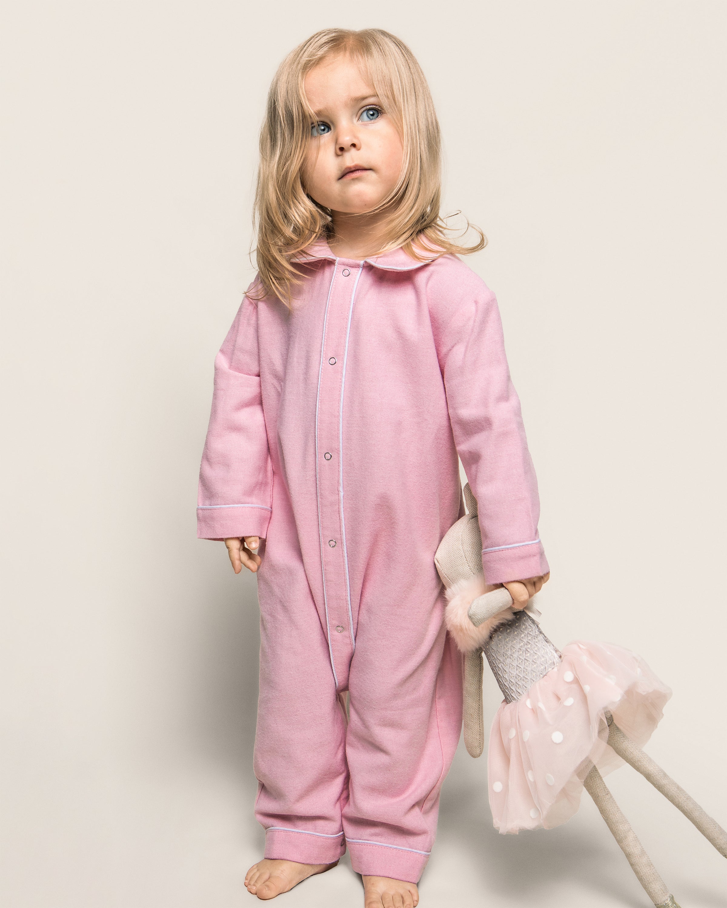 A young child with long blonde hair wears Petite Plumes Babys Flannel Cambridge Romper in Pink. They hold a stuffed toy bunny in a pink tutu against a plain, light-colored background that enhances the cozy feel of the childrens sleepwear.