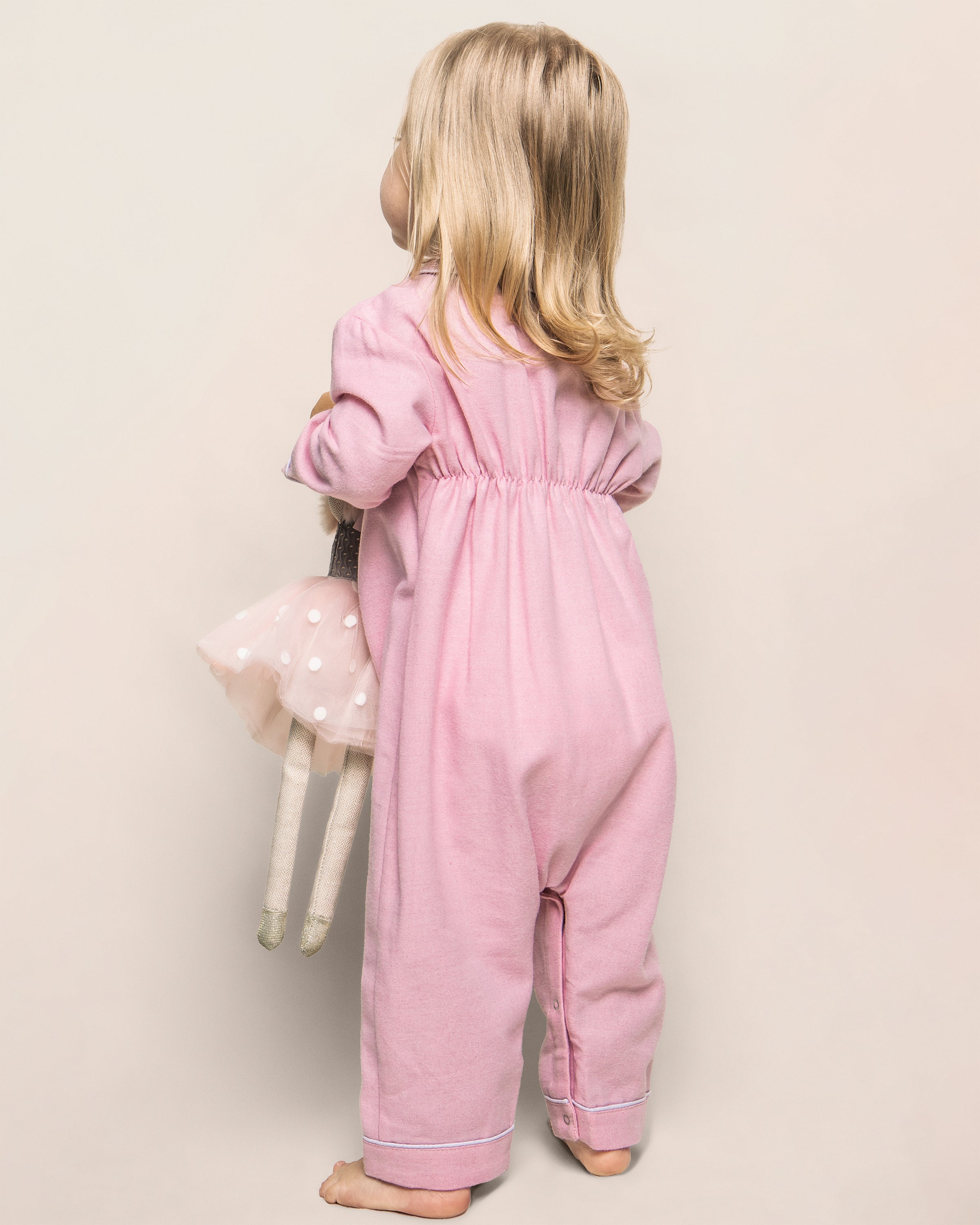 A toddler with blond hair stands barefoot on a plain background, wearing Petite Plumes Babys Flannel Cambridge Romper in Pink. She holds a doll dressed in a pink tutu adorned with white polka dots, her head turned slightly to the side.