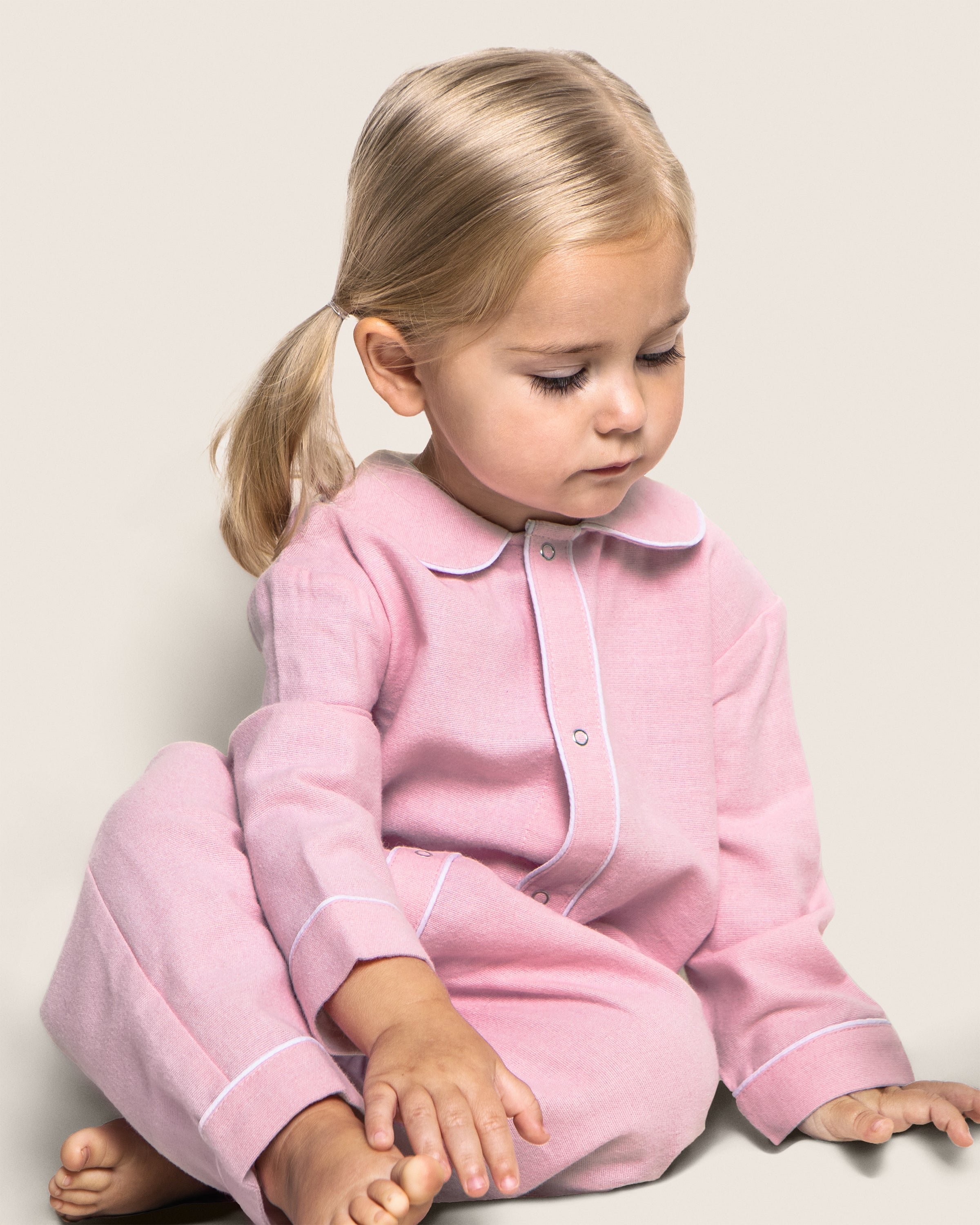 A blonde toddler sits on the floor wearing Petite Plumes Babys Flannel Cambridge Romper in Pink, crafted with brushed cotton flannel and white trim for comfort and safety. The childs head is slightly bowed, calm and focused against a soft, neutral background.