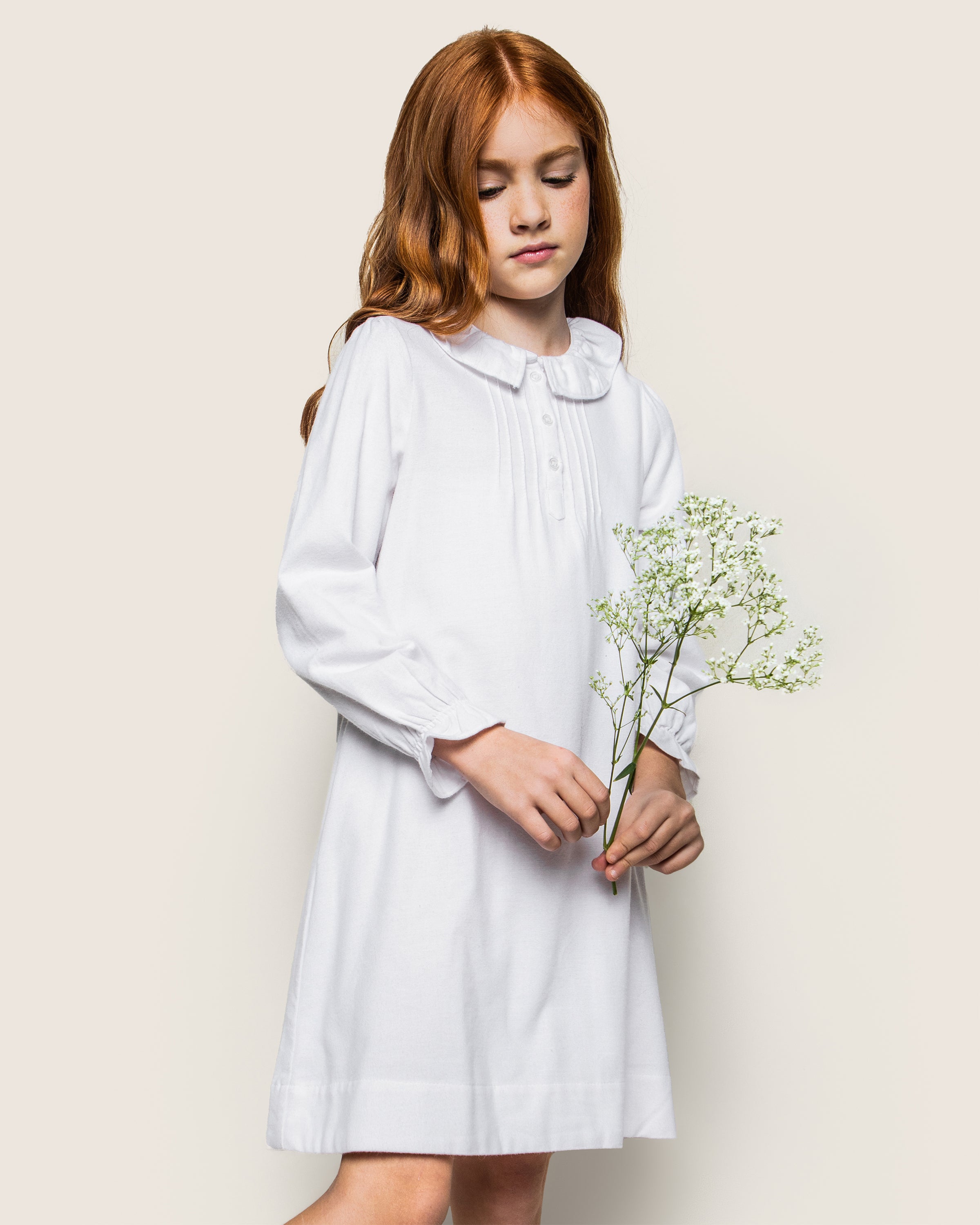 A young girl with long red hair wears Petite Plumes Girls Flannel Victoria Nightgown in white, featuring a Peter Pan collar. She stands against a plain background, holding a small bouquet of white flowers.