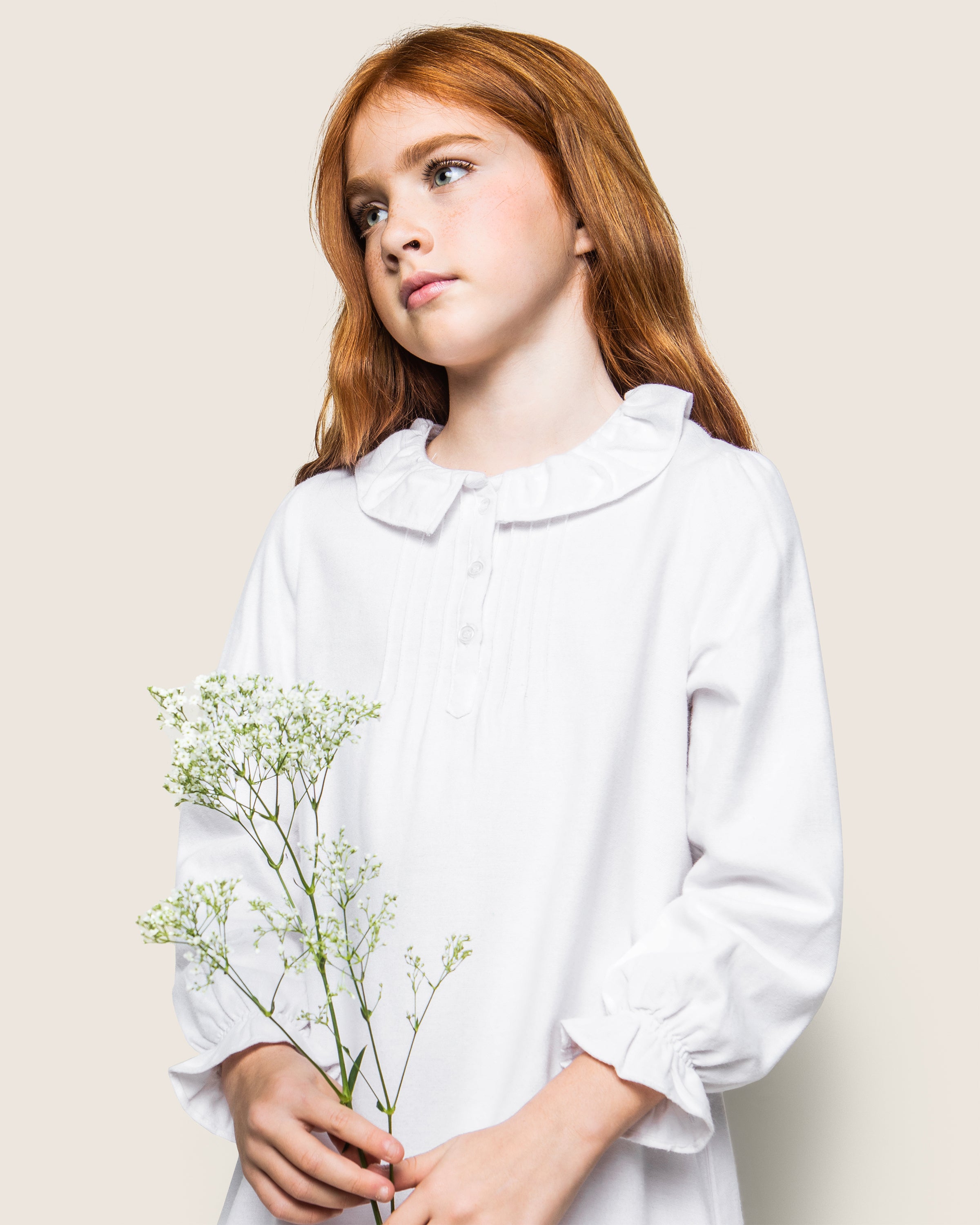 A red-haired girl in Petite Plumes Girls Flannel Victoria Nightgown in White, featuring a ruffled collar, holds a small bouquet of white flowers. She gazes to the side against a plain beige background, exuding vintage sleepwear elegance.