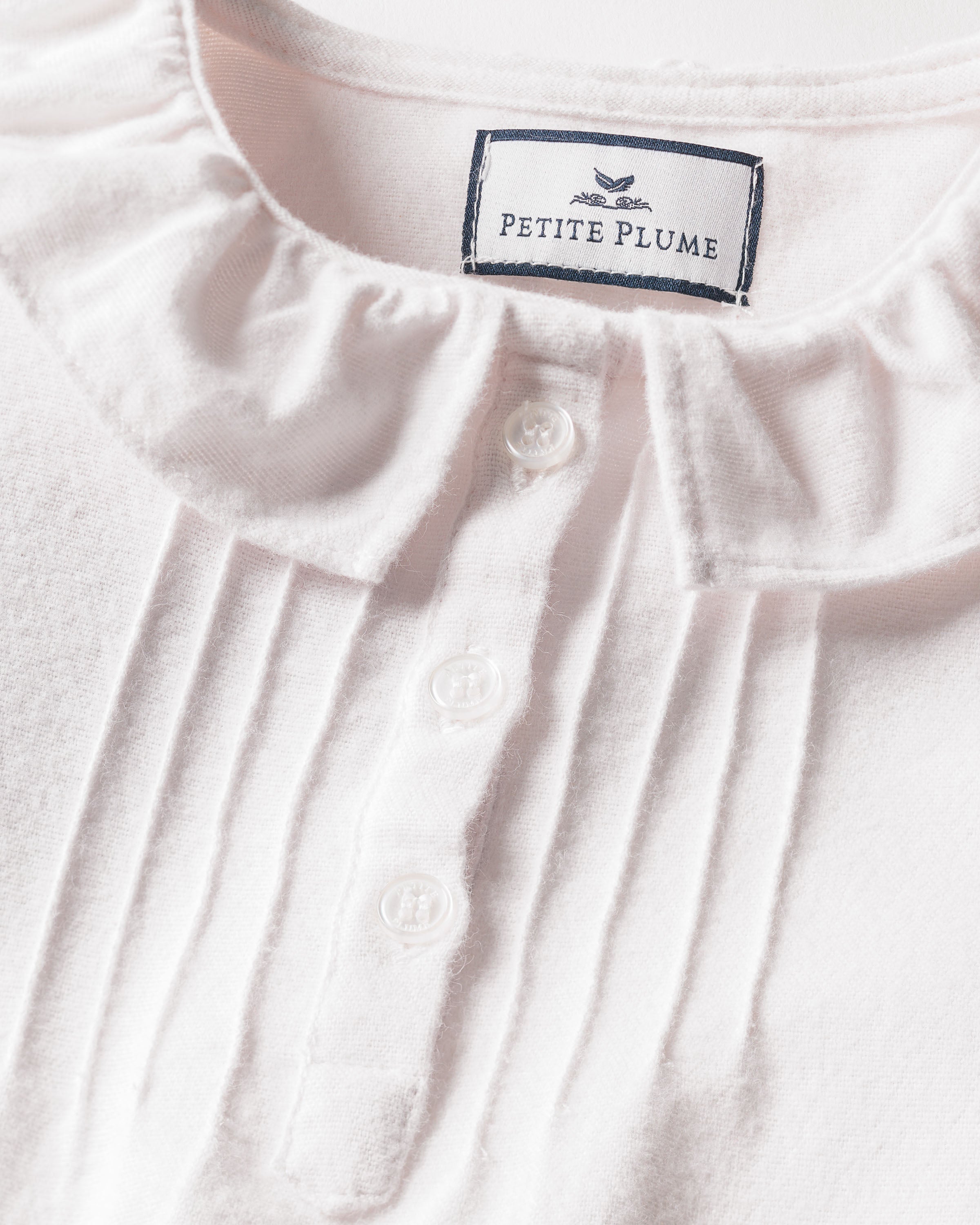 Close-up of a white flannel nightgown called the Girls Flannel Victoria by Petite Plume, featuring a ruffled collar and a four-button front placket. The fabric looks soft and textured, with the brand label sewn inside the collar.