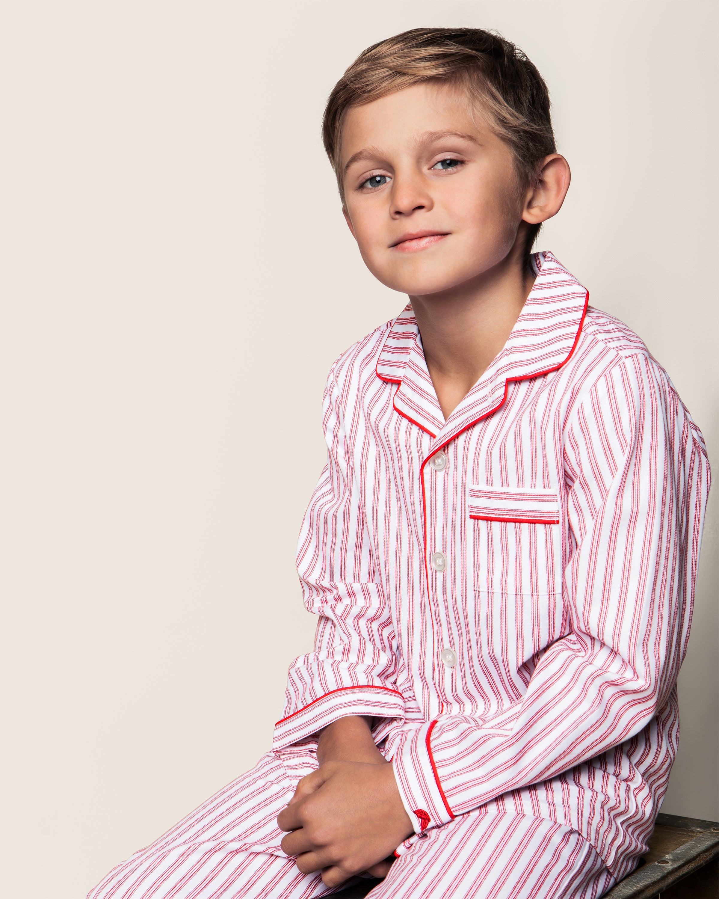 A young boy sits on a wooden stool wearing Petite Plumes Kids Twill Pajama Set in Antique Red Ticking. His short, light brown hair and slight smile face the camera against a plain, light background.