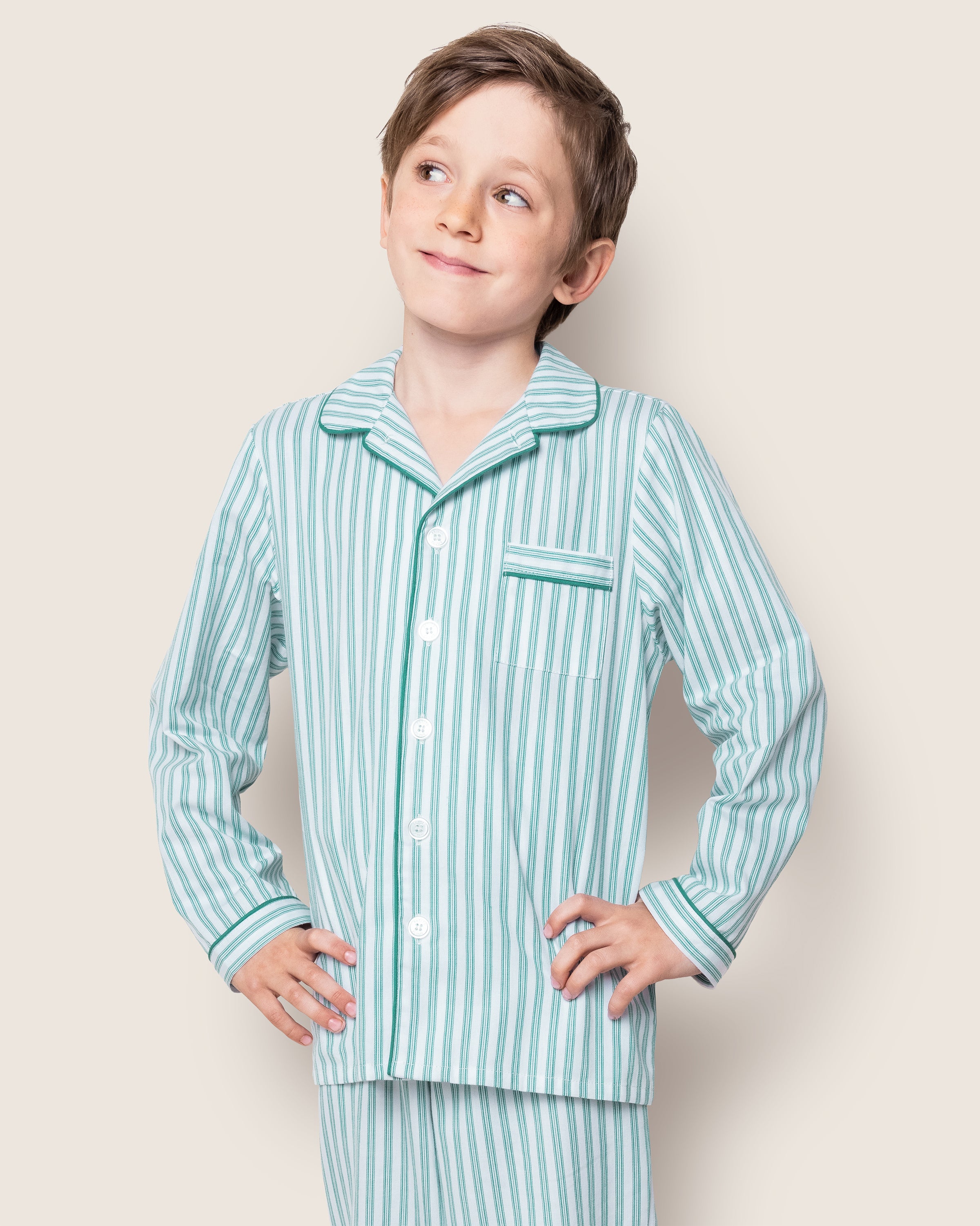 A child in an Emerald Ticking twill pajama set by Petite Plume, featuring buttons and a chest pocket, smiles with hands on hips against a light, neutral background.