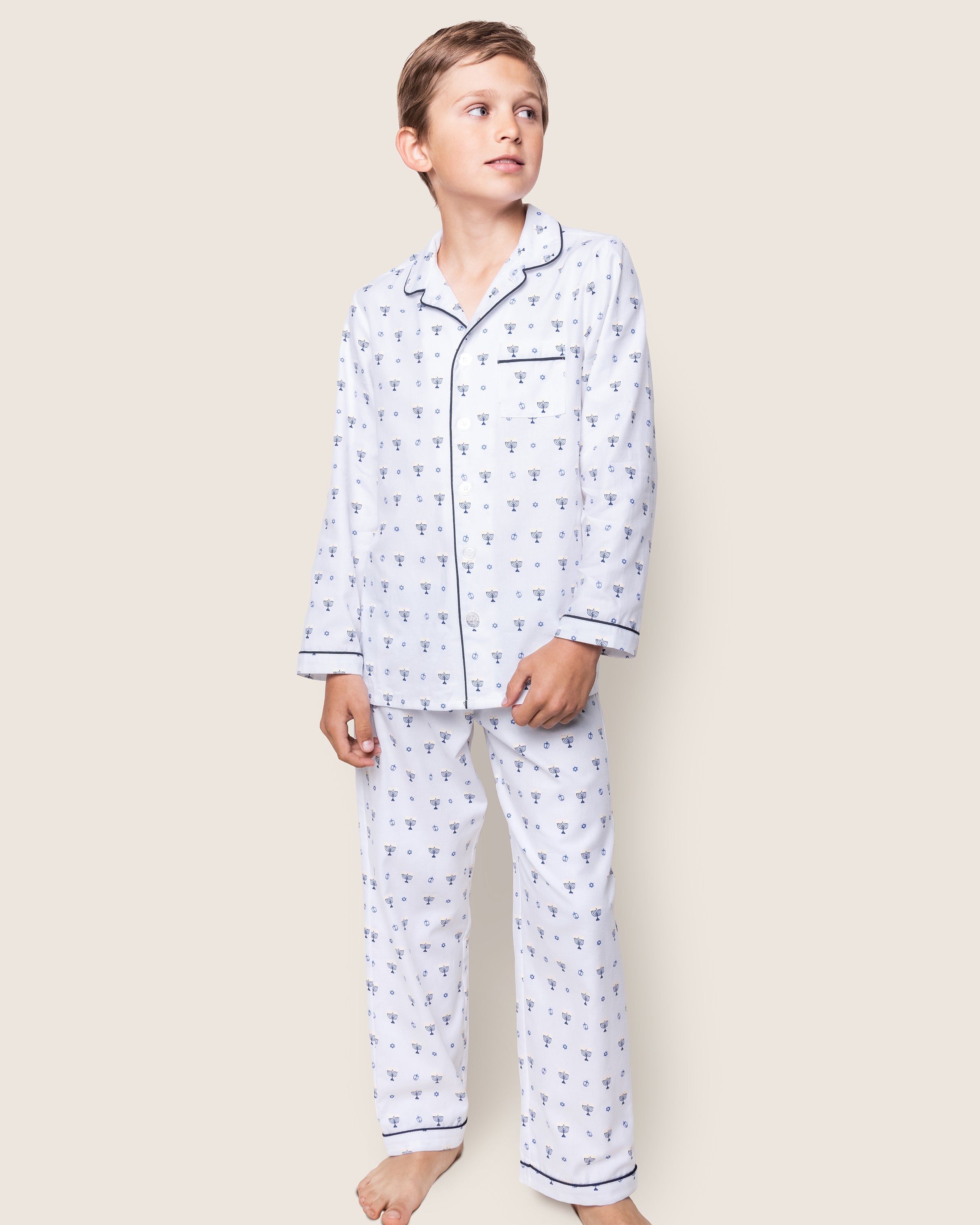 A young boy stands barefoot in Petite Plumes Kids Twill Pajama Set, Happy Hanukkah. The collared long-sleeve top and matching pants exude comfort, as he gazes upward against a plain beige background.