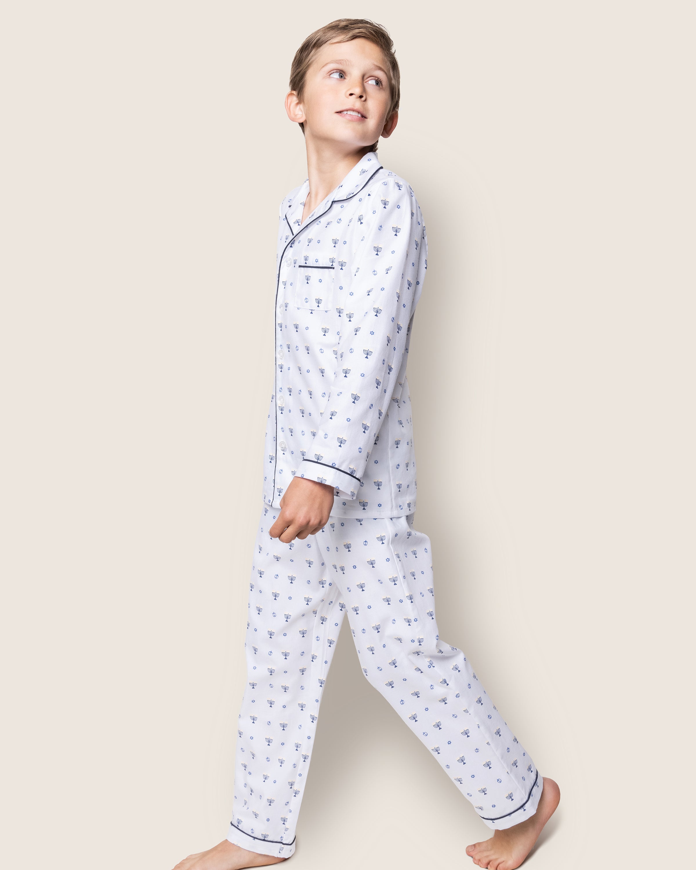 A young boy wears the Petite Plume Kids Twill Pajama Set in Happy Hanukkah, featuring a button-up top and matching pants with small blue patterns. He looks up as he walks barefoot against a plain, light background.