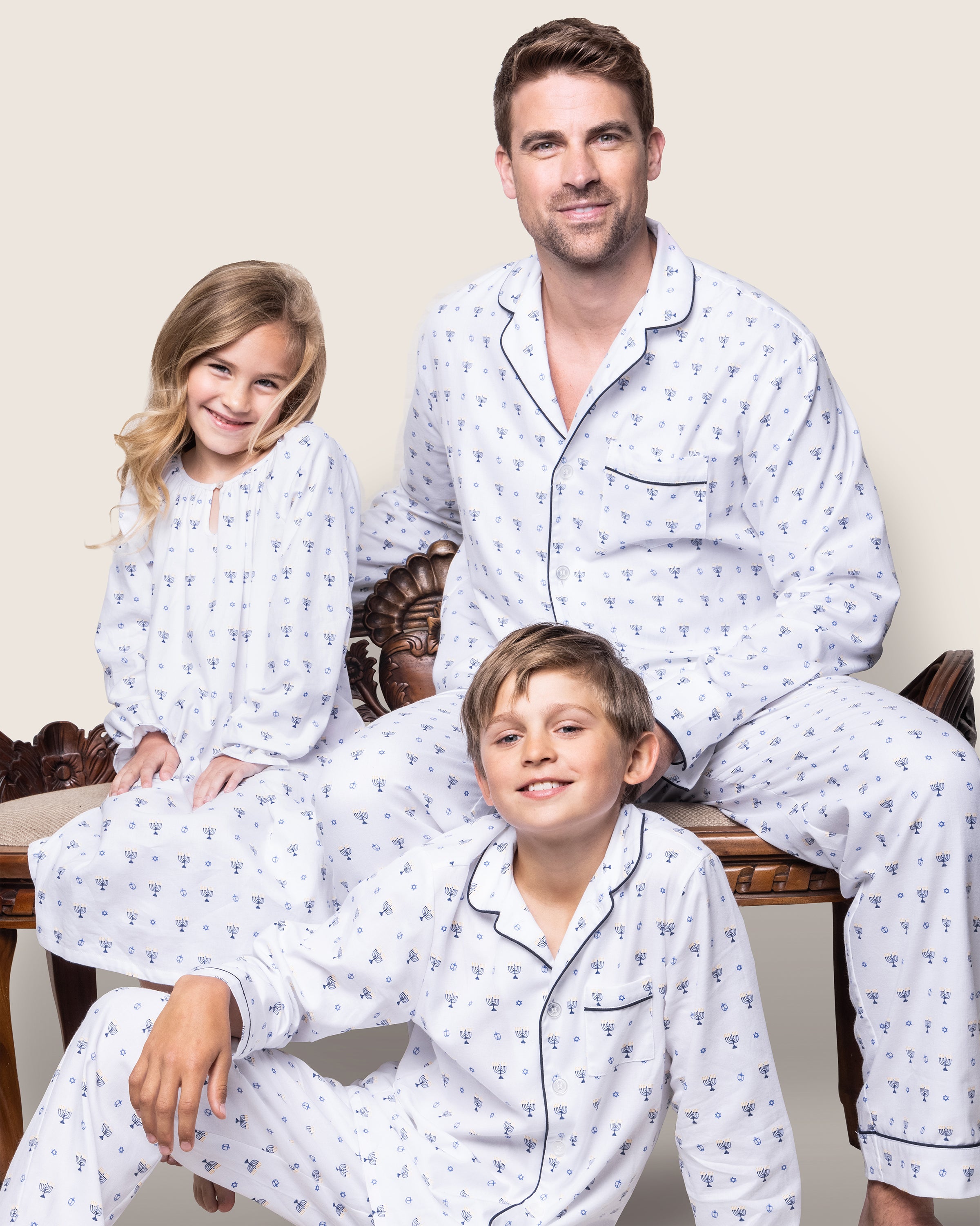 A man and two children, all in matching Petite Plume Kids Twill Pajama Sets in Happy Hanukkah, are seated on a wooden bench. The girl and boy wear white pajamas with a blue pattern, smiling brightly beside the gentle-faced man.