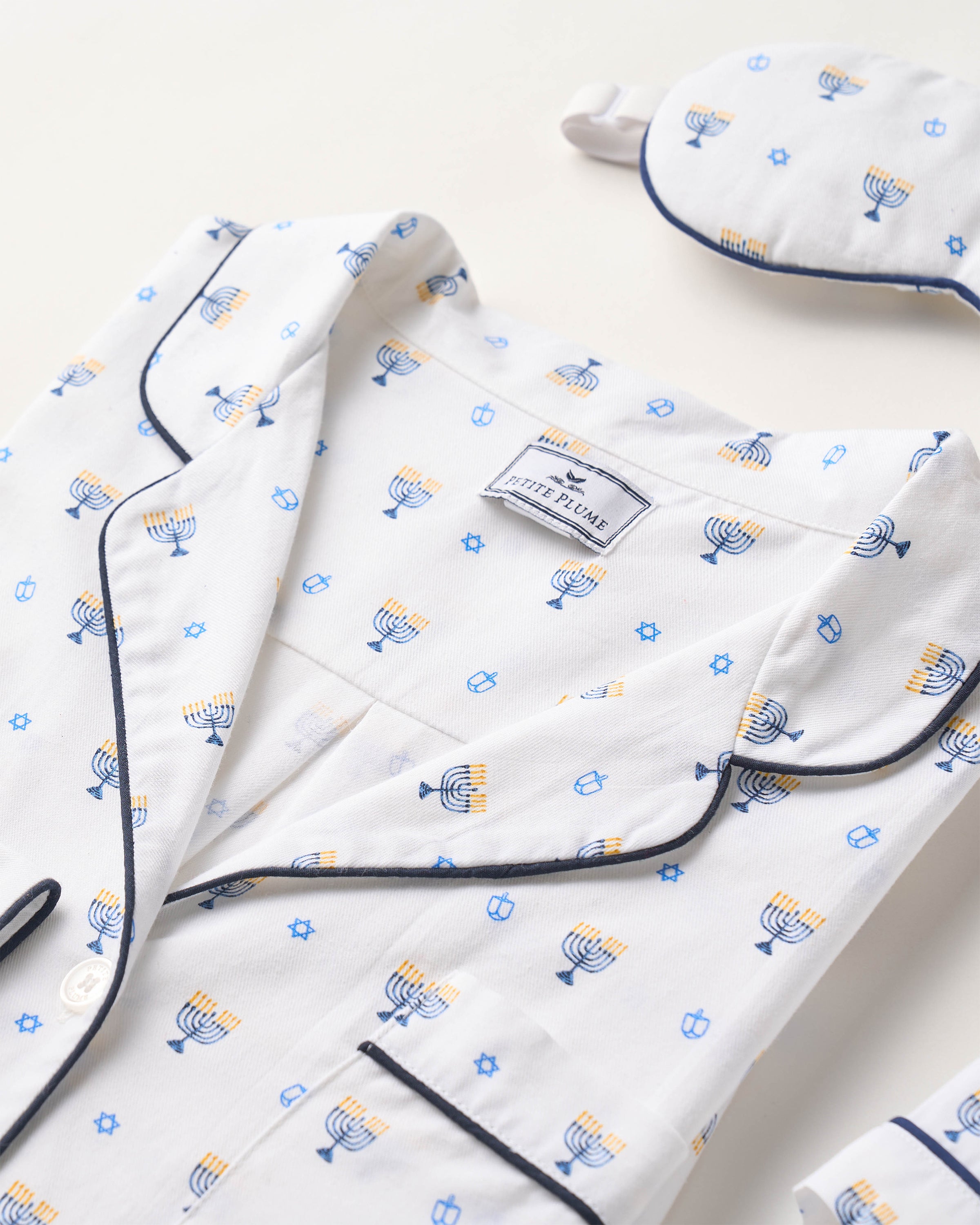 The Kids Twill Pajama Set in Happy Hanukkah by Petite Plume boasts a white design with blue and yellow menorah, dreidel, and Star of David patterns. It features blue piping on the shirt and includes a matching eye mask, all made in moisture-wicking fabric for cozy nights.