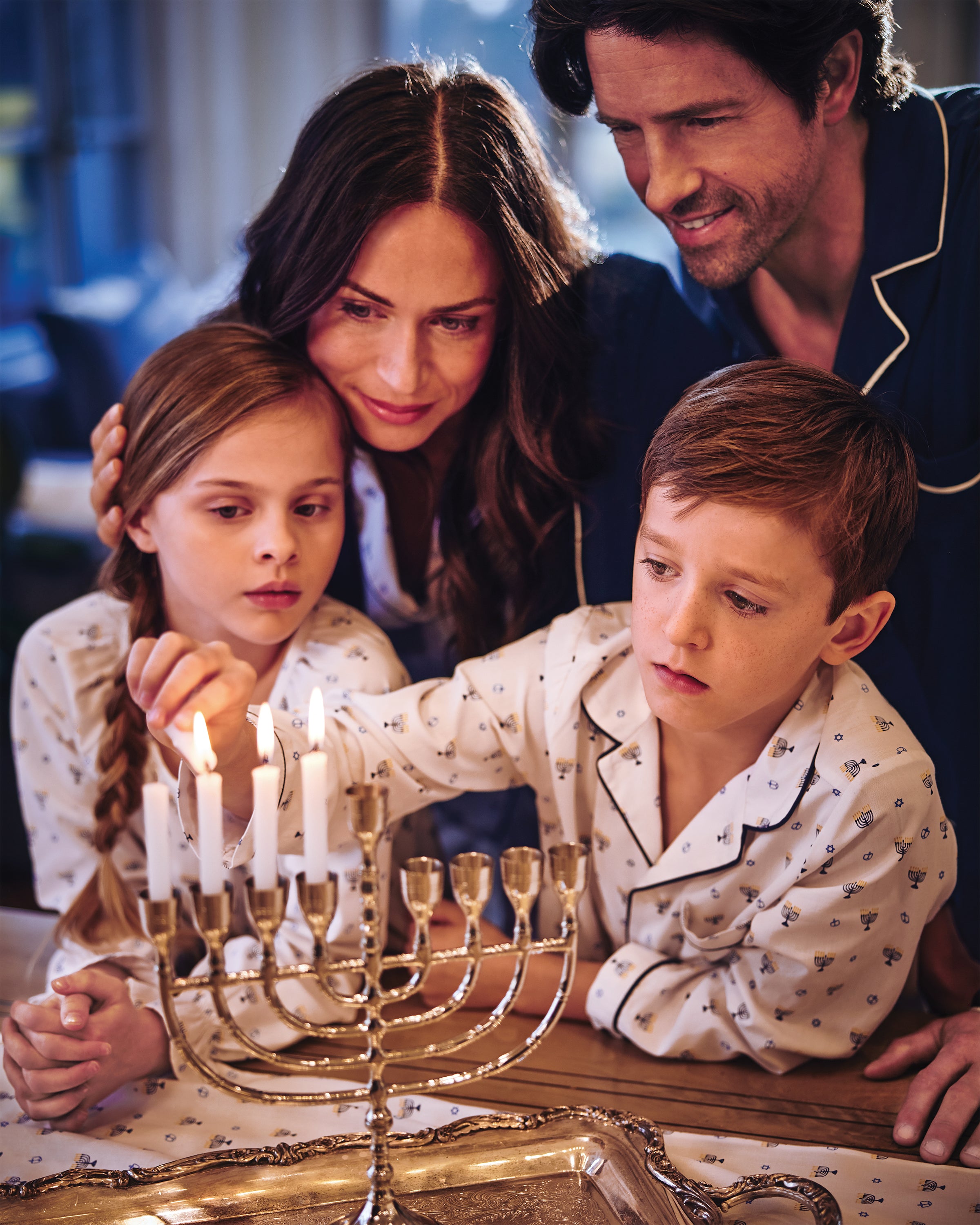 A family of four in Petite Plumes Kids Twill Pajama Set in Happy Hanukkah gathers by a lit menorah. The mother embraces the braided-hair girl while the father stands behind them, as the boy reaches to light a candle, creating a warm and cozy scene.