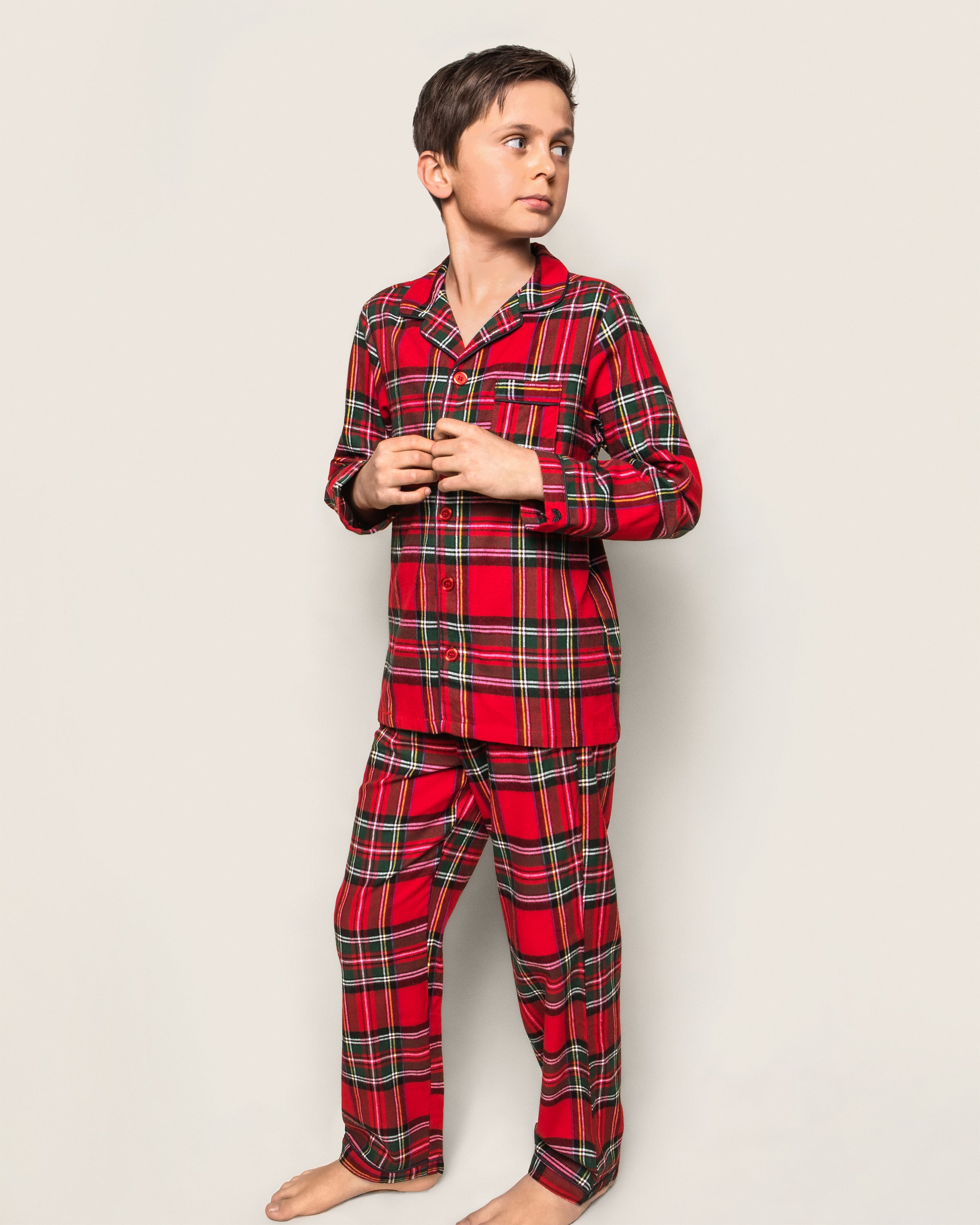 A boy in Petite Plumes Kids Brushed Cotton Pajama Set in Imperial Tartan stands barefoot against a plain background, looking to his right.