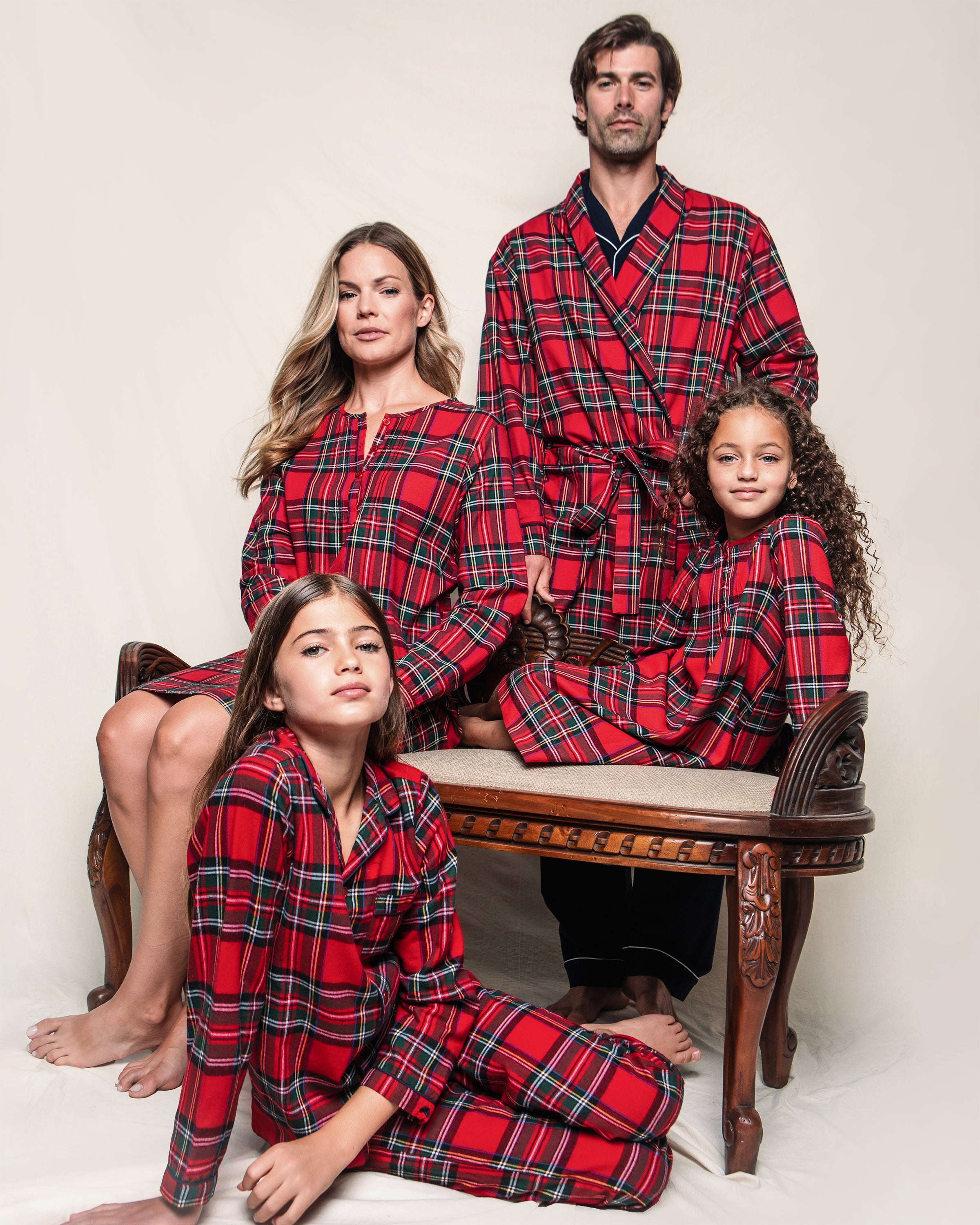 A family of four poses in matching Imperial Tartan pajamas. The father stands behind a wooden bench, the mother wears a Womens Brushed Cotton Beatrice Nightgown from Petite Plume as she sits next to him, with two young girls leaning against the bench. They all face the camera against a plain backdrop.