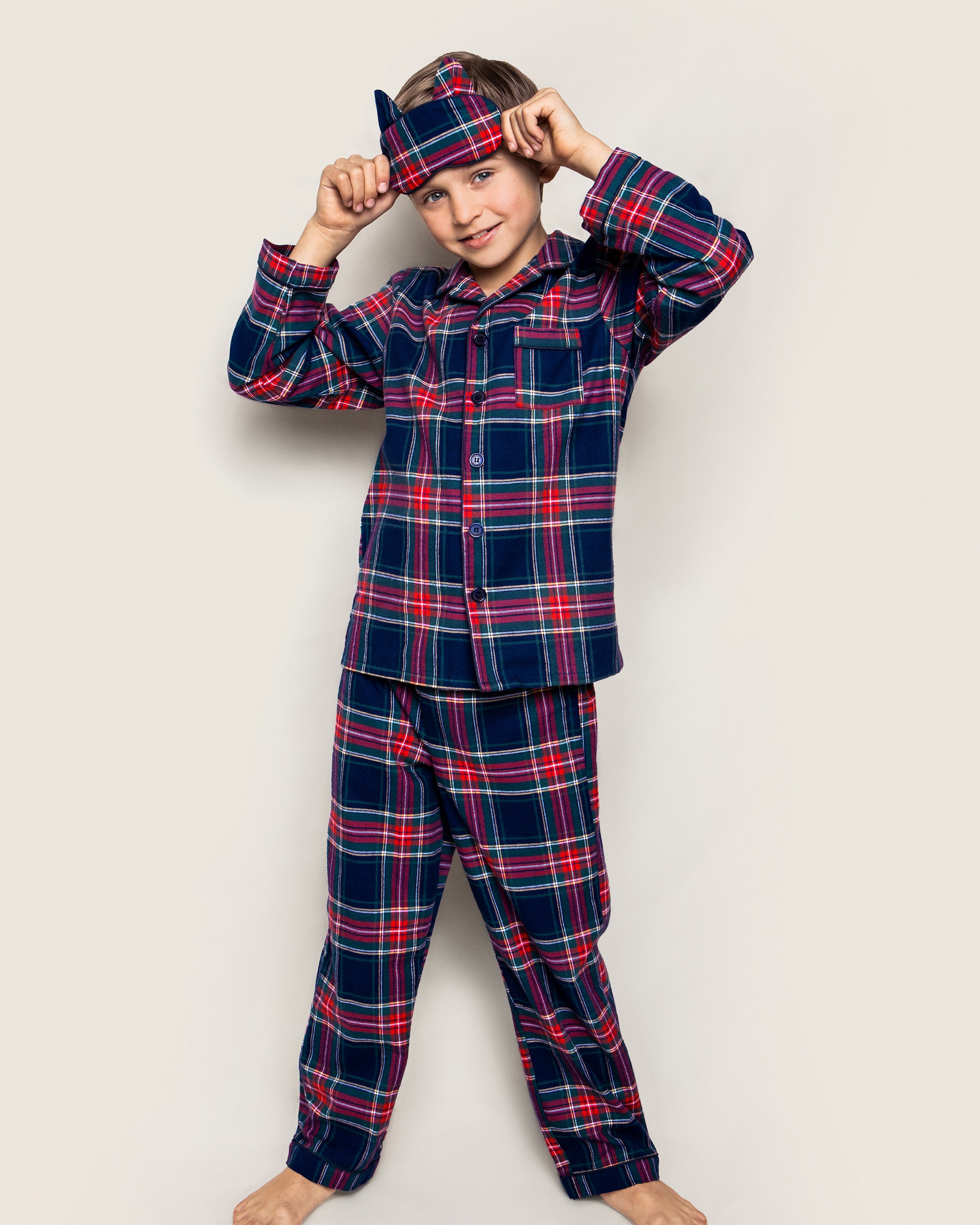 A boy in a Petite Plume Kids Brushed Cotton Pajama Set in Windsor Tartan, paired with a matching sleep mask, stands barefoot against a plain background, adjusting the mask with a smile.