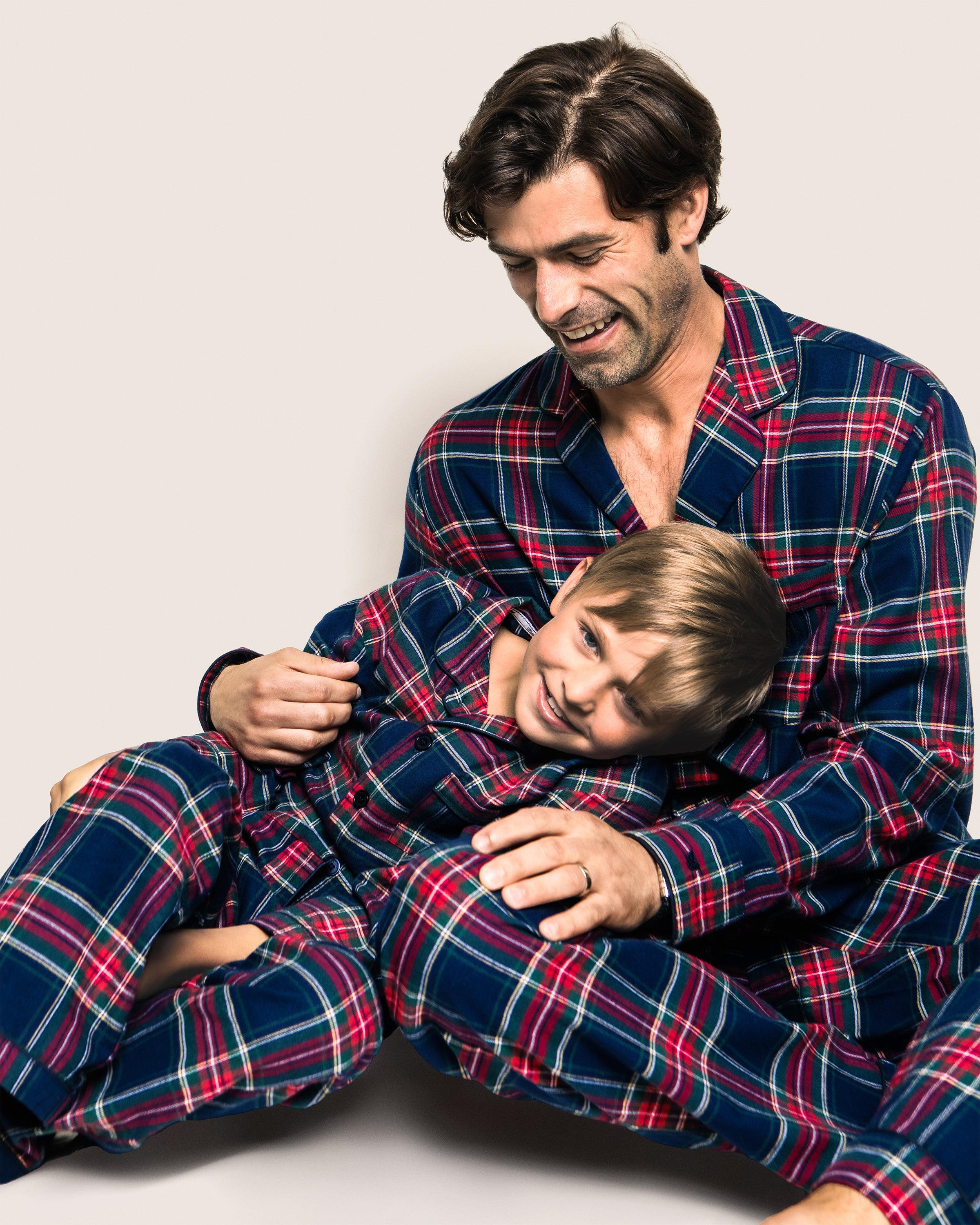 A man and young boy share a cozy moment in Petite Plumes Kids Brushed Cotton Pajama Set in Windsor Tartan, with the man smiling and holding the content boy against a plain, light backdrop.