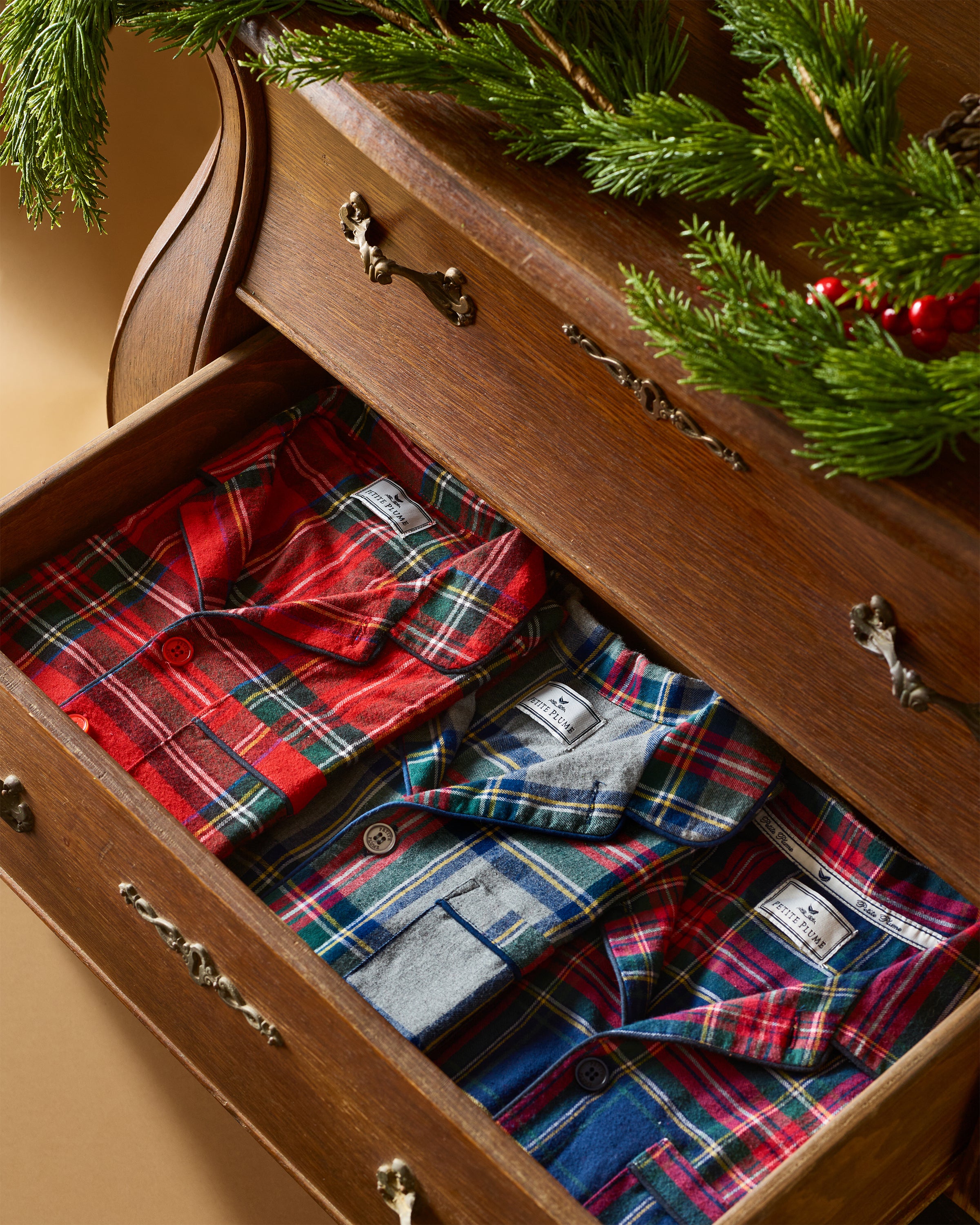 An open wooden drawer reveals three folded Petite Plume Kids Brushed Cotton Pajama Sets in Windsor Tartan, featuring red, green, and blue patterns. Above the drawer, a festive garland with greenery and red berries adorns the dresser, crafting a holiday ambiance.