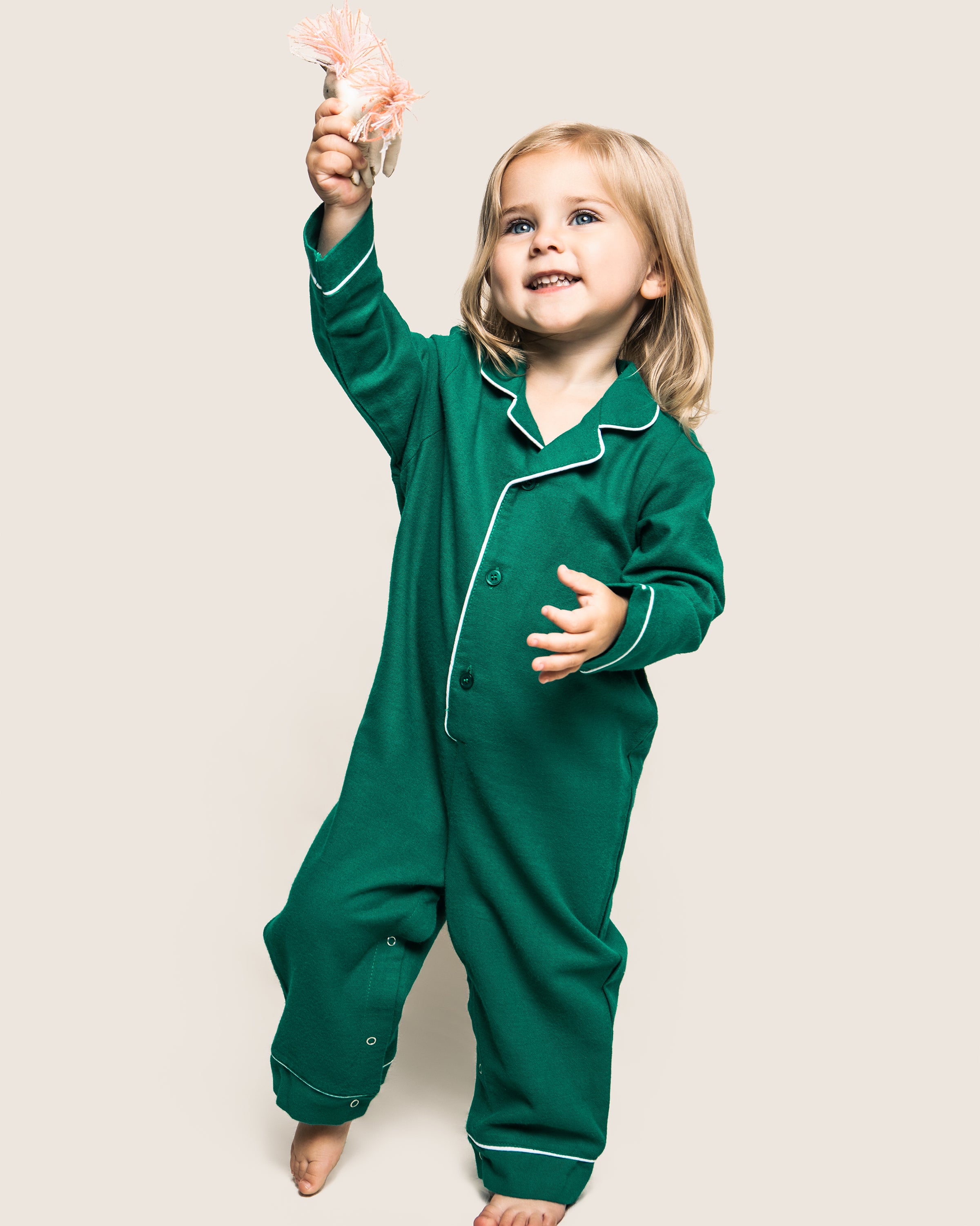 A joyful blonde child stands barefoot against a plain background, holding a plush toy while wearing Petite Plumes Babys Flannel Romper in Forest Green with White Piping.