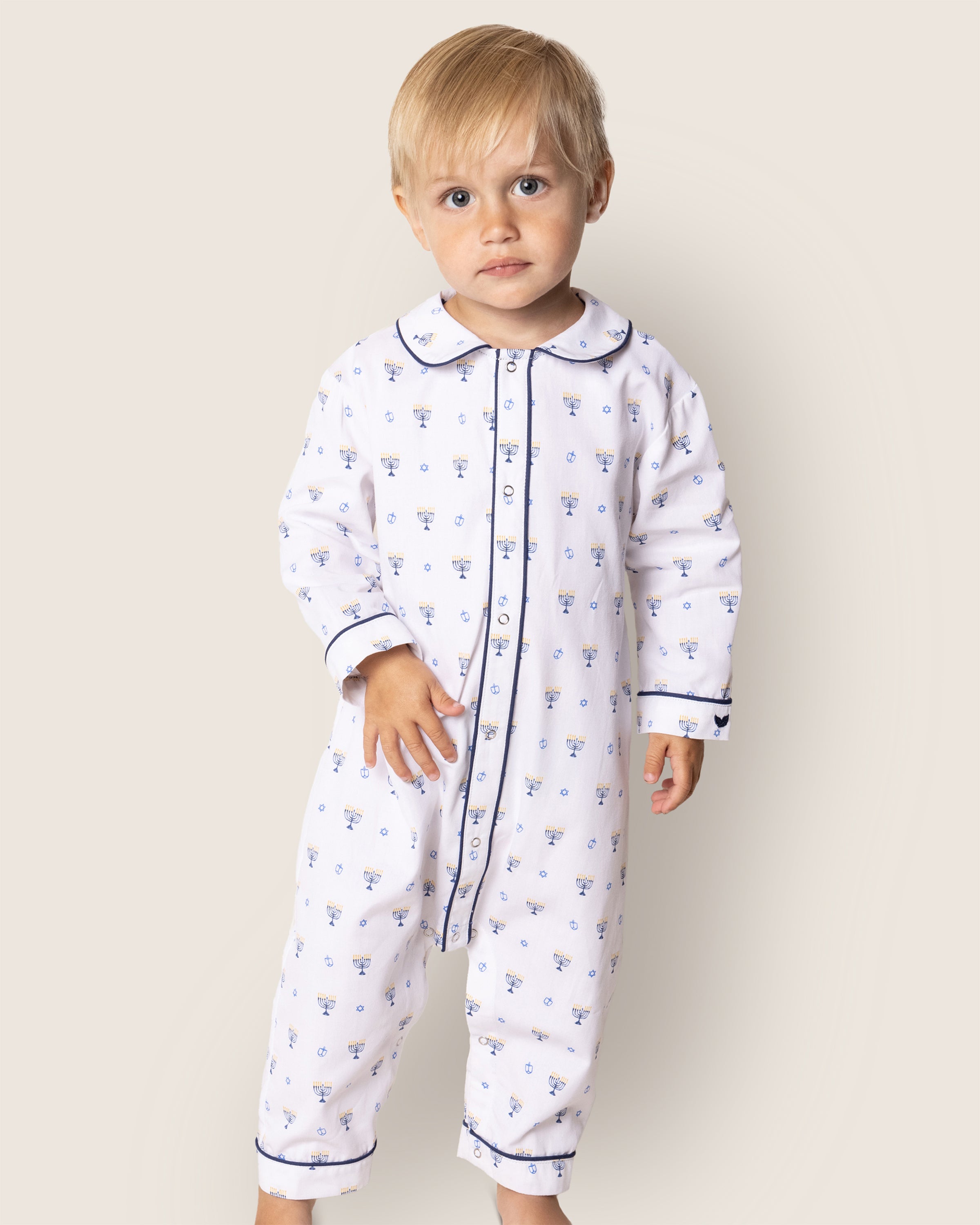 A young child with blonde hair wears Petite Plumes Babys Twill Cambridge Romper in Happy Hanukkah, featuring a blue floral pattern with dark trim and a collared button-down front. Made from chemical-free materials, the romper is set against an off-white background as the child looks at the camera.