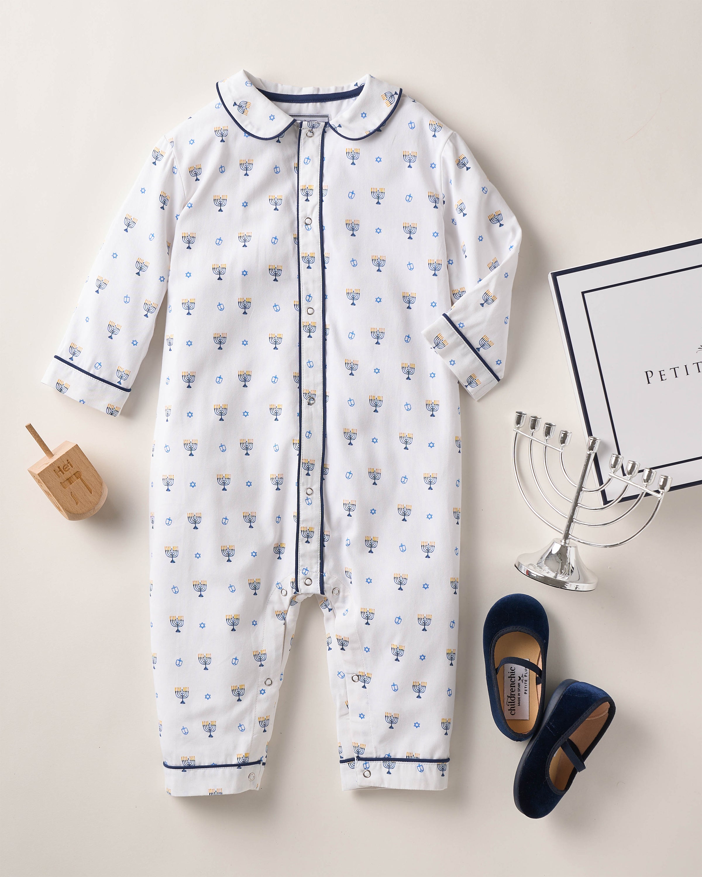 The Petite Plume Babys Twill Cambridge Romper in Happy Hanukkah design, featuring blue and yellow floral patterns, a menorah, dreidel, and blue baby shoes on a flame-retardant fabric, rests on a light surface. Perfect for Hanukkah celebrations, it comes in a decorative box.