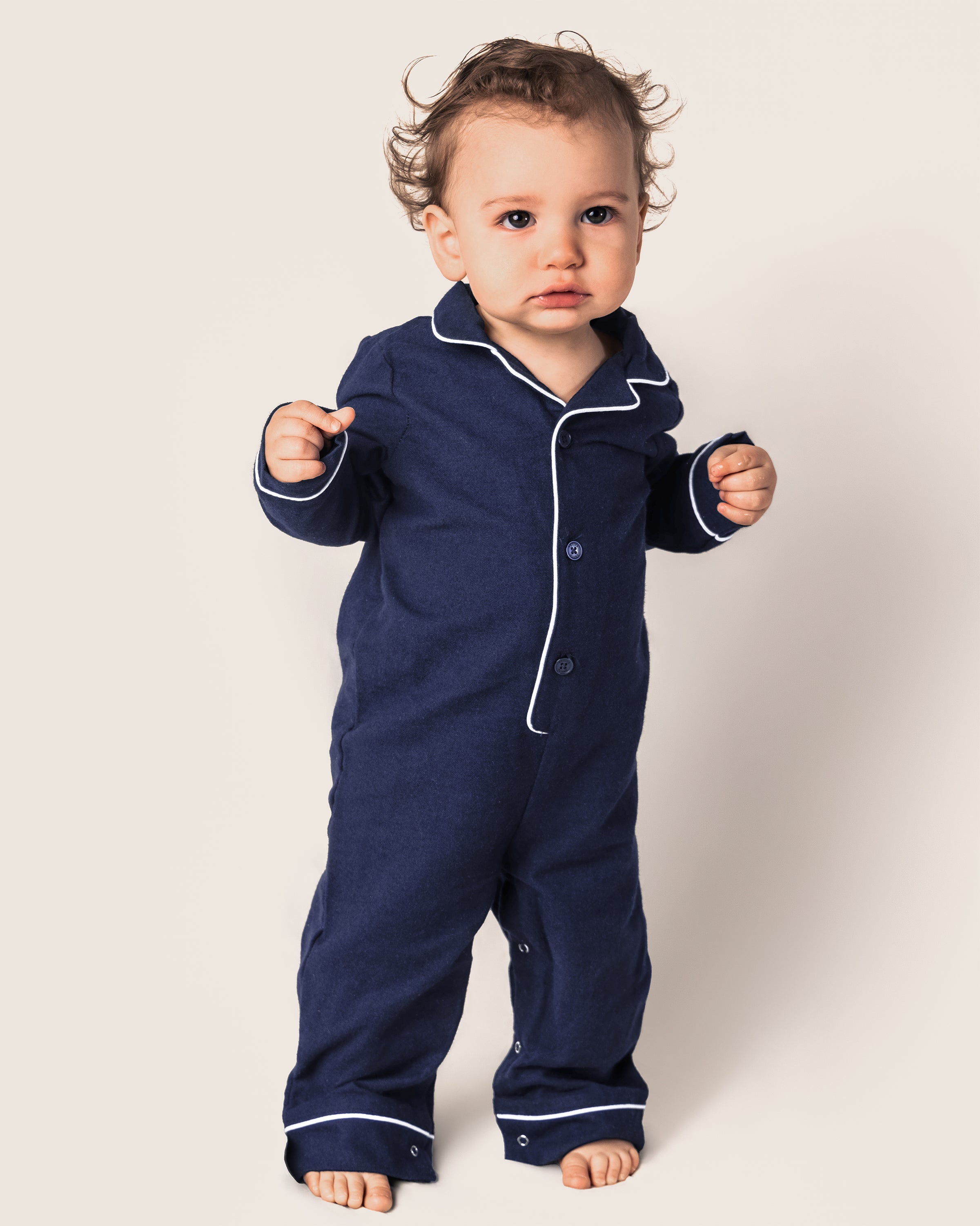 A toddler with curly hair and an inquisitive look stands barefoot, wearing the Petite Plume Babys Flannel Romper in navy with white piping, against a plain light background.