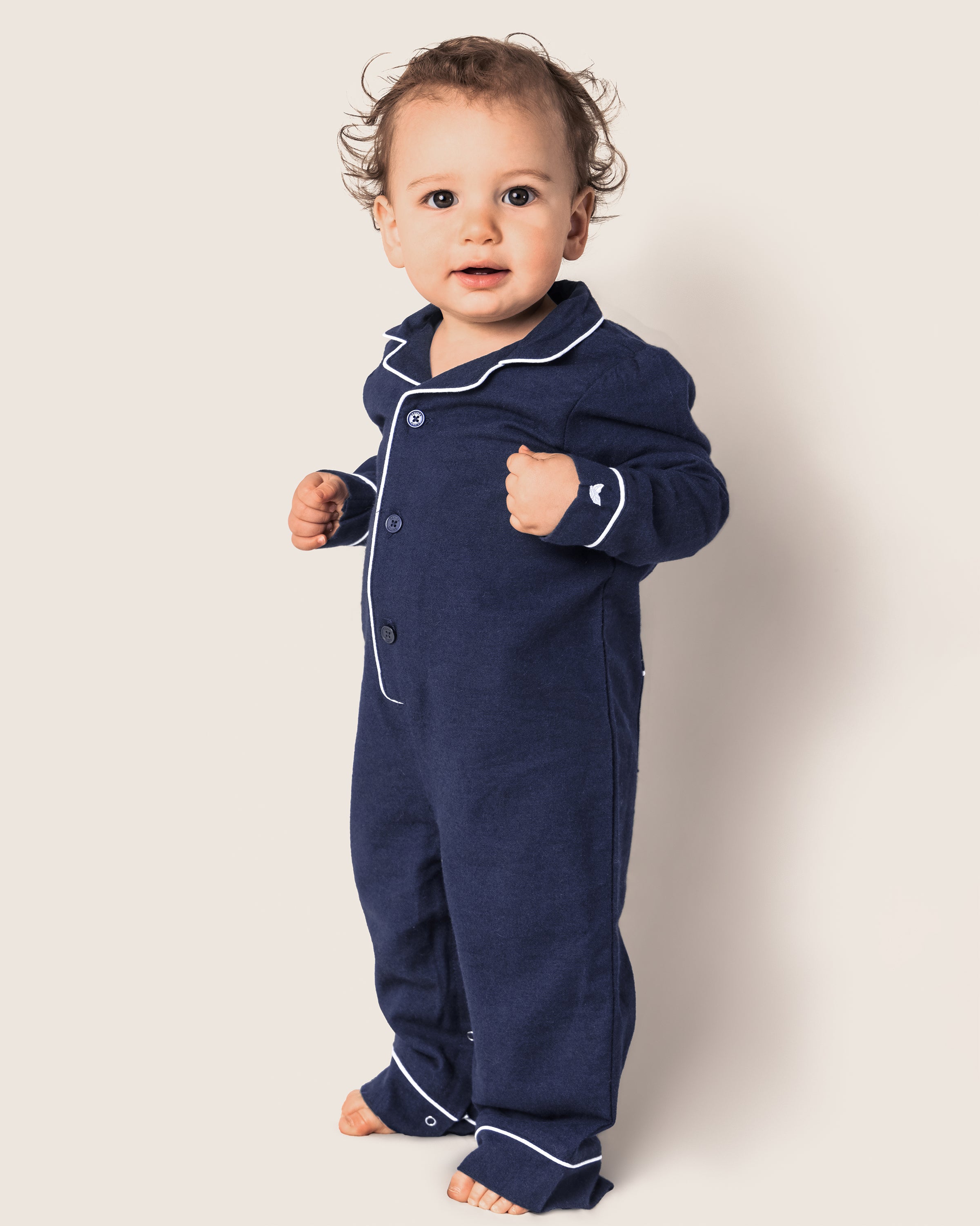 A toddler with curly hair stands mid-step, hands slightly clenched and wearing Petite Plumes Babys Flannel Romper in Navy with White Piping, facing the camera against a light background that enhances the charm of the cozy sleepwear moment.