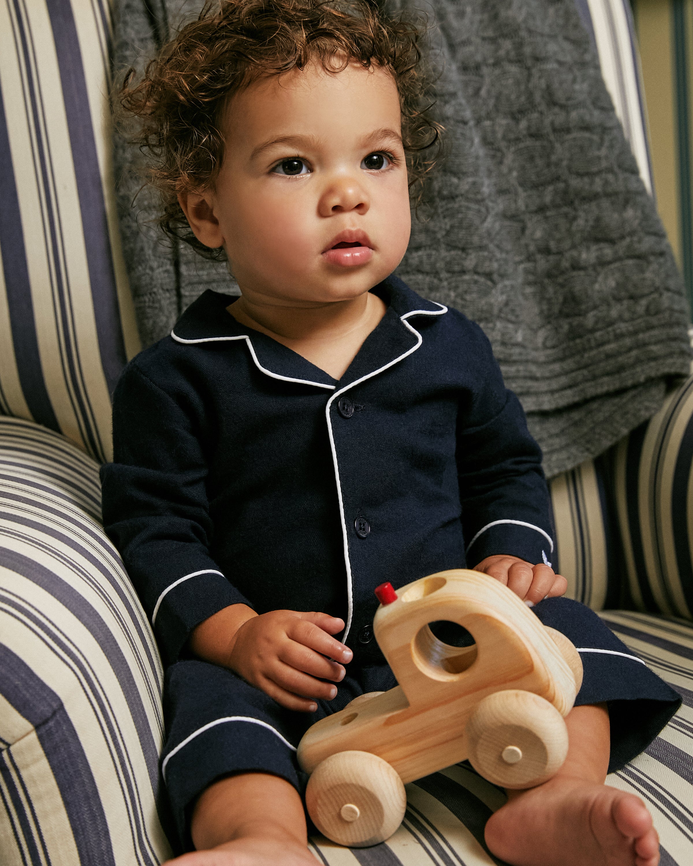 A toddler with curly hair sits on a striped chair, wearing Petite Plumes Babys Flannel Romper in Navy with White Piping. The child holds a wooden toy car and looks ahead curiously. A textured gray blanket is draped over the chair.