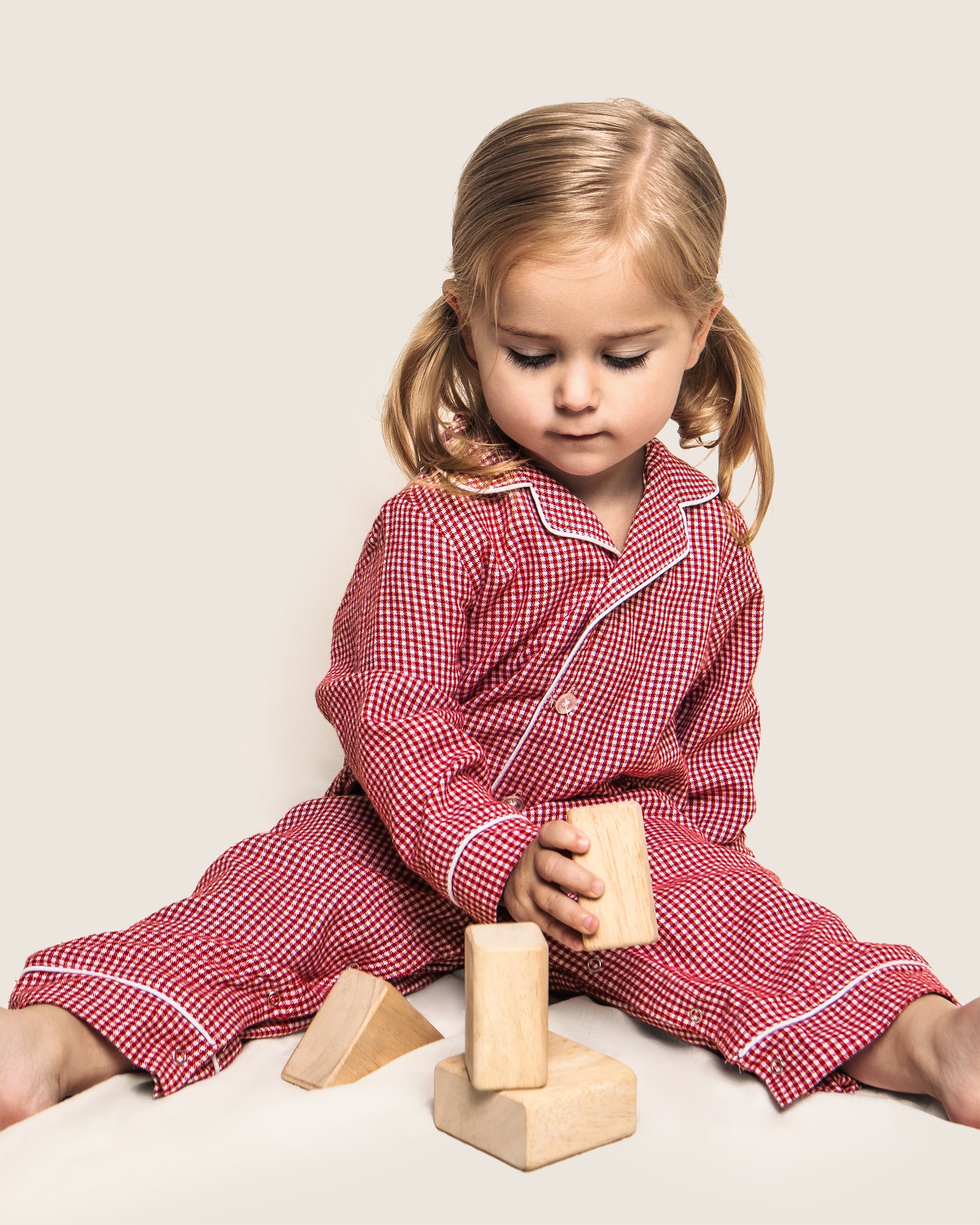 A young child with blonde hair sits on the floor in a Petite Plume Babys Flannel Romper in Red Mini Gingham, intently stacking wooden blocks into a small tower against a plain, light-colored background.